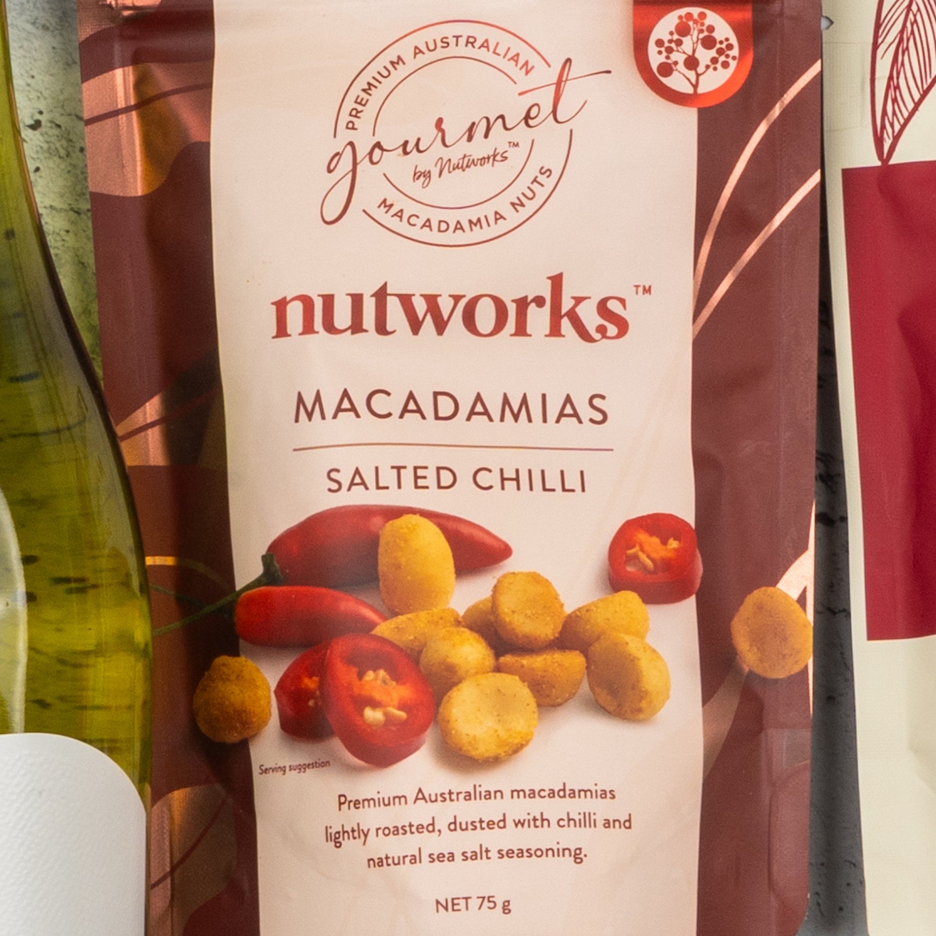 A Gourmet Getaway Gift Hamper by The Hamper Boutique Co, featuring a bag of macadamias with salted chilli flavor, its packaging displaying images of nuts and red chili peppers, placed partially beside a wine bottle.