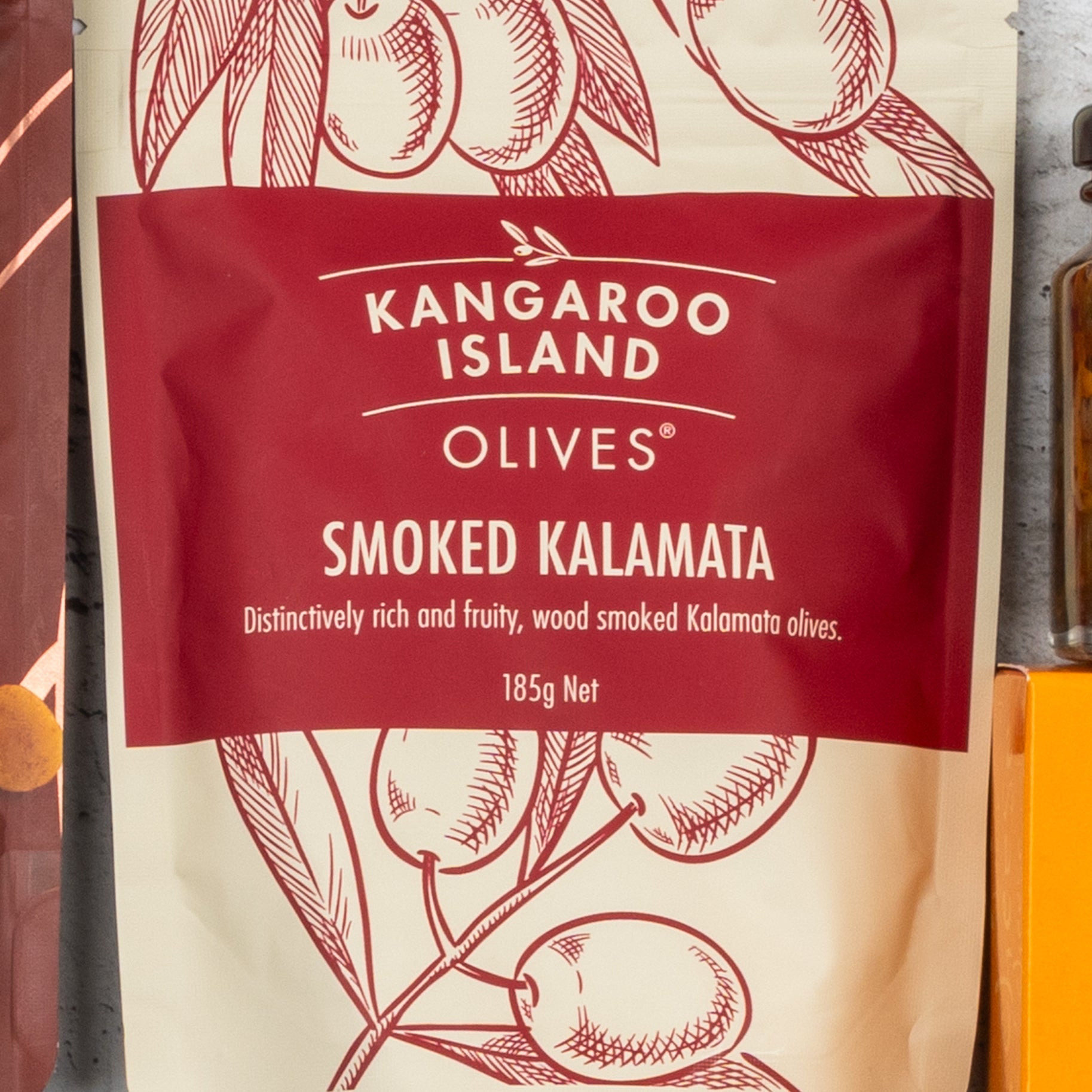 A 185g red and cream packet of Smoked Kalamata Olives from The Hamper Boutique Co’s Gourmet Getaway Gift Hamper, featuring an illustrated olive branch and described as distinctively rich, fruity, and wood smoked.
