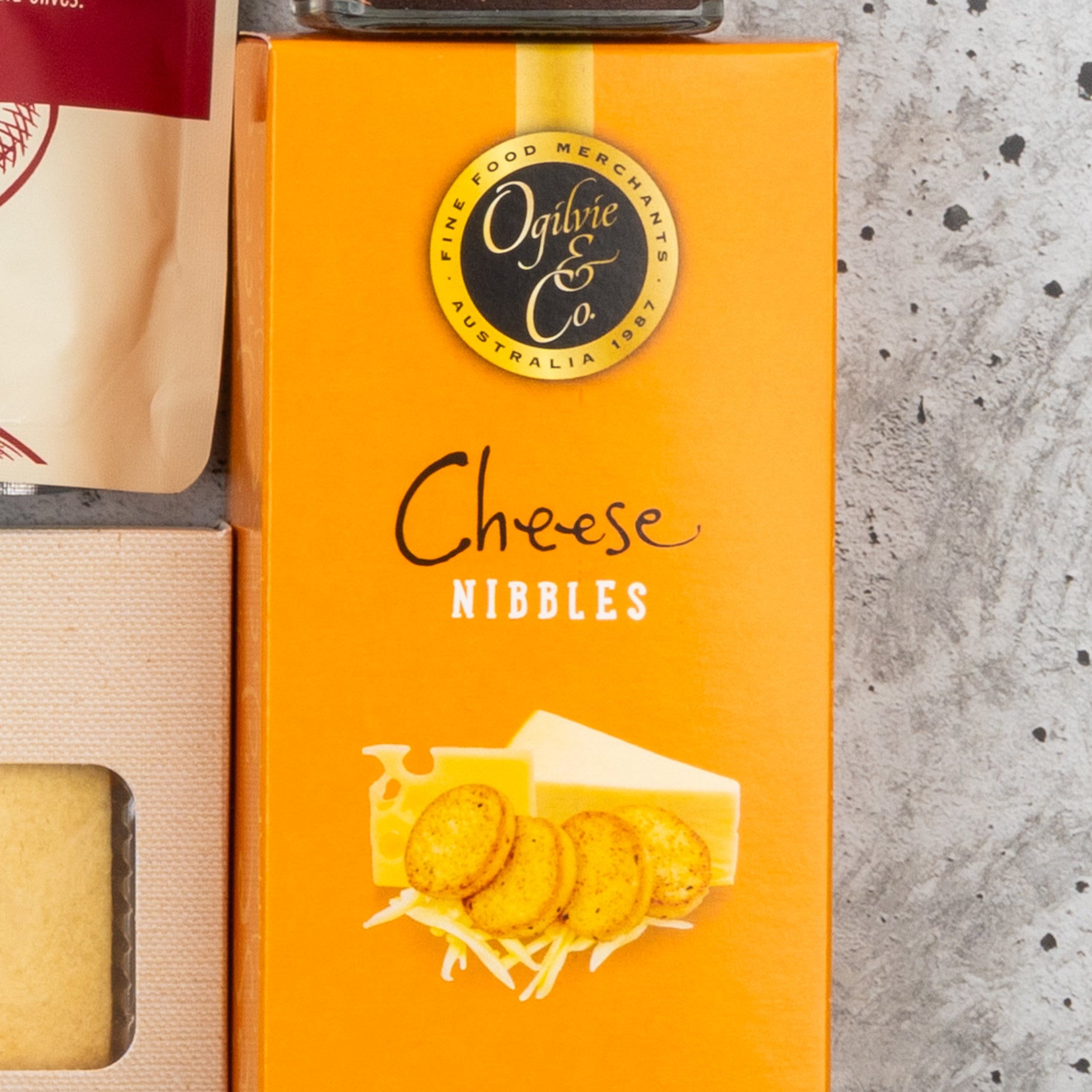 A Gourmet Getaway Gift Hamper by The Hamper Boutique Co in an orange box labeled Cheese Nibbles with images of cheese and crackers, plus other packaged foods, is displayed on a speckled gray surface.