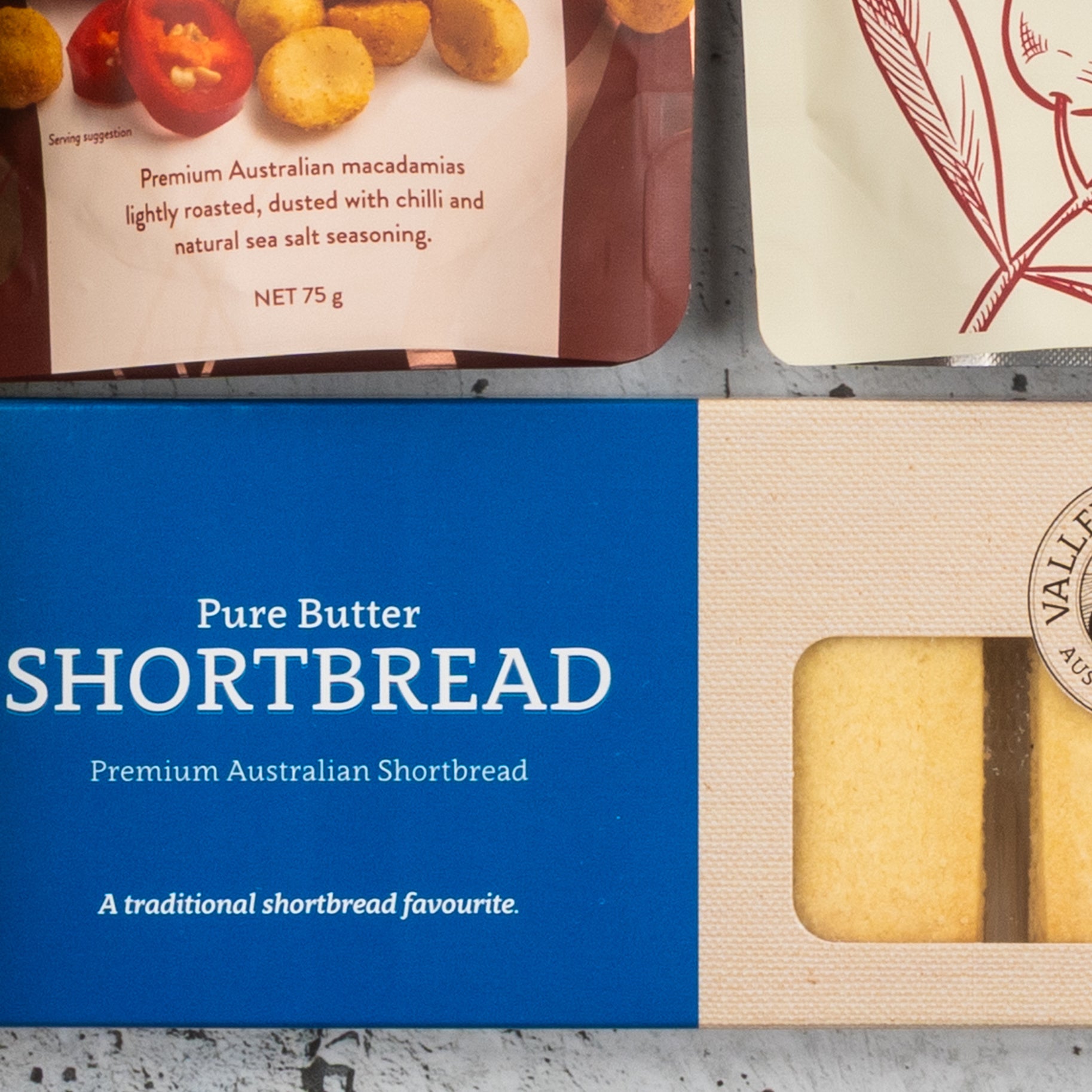 A box of pure butter shortbread with a blue label and a bag of chili sea salt roasted macadamias are shown. At the top right, part of another beige and red item can be seen from The Hamper Boutique Co's Gourmet Getaway Gift Hamper.