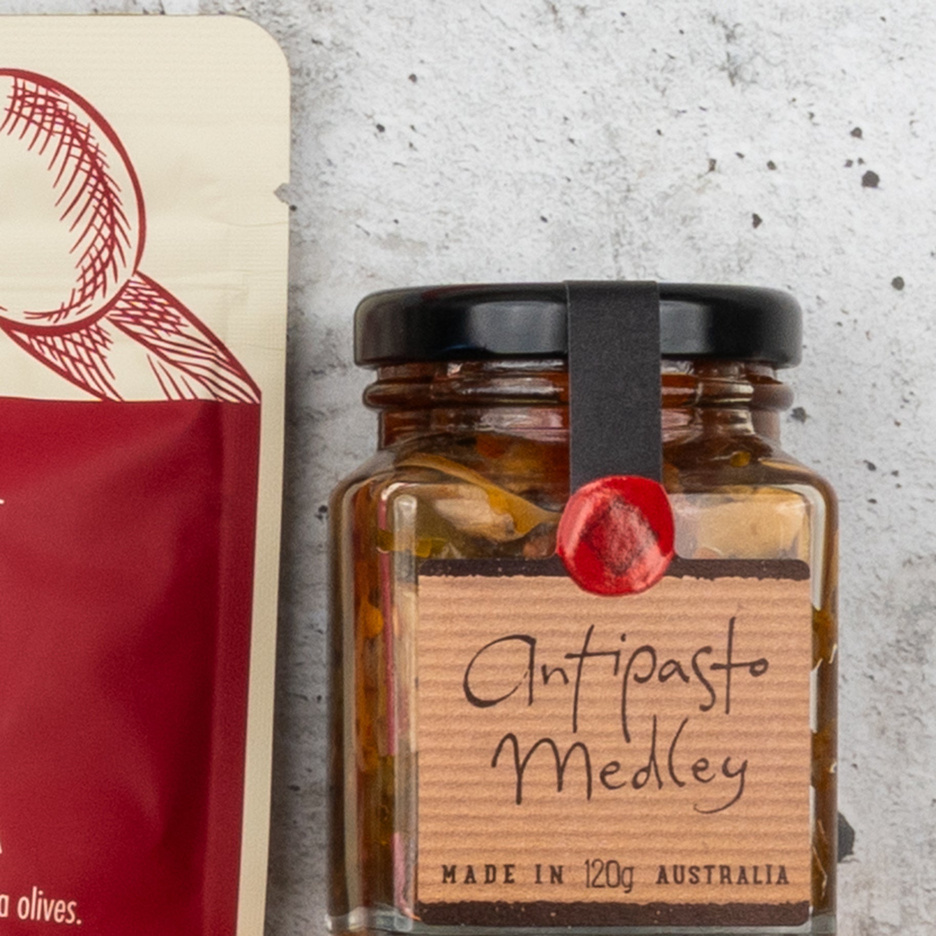 A close-up of a Gourmet Getaway Gift Hamper from The Hamper Boutique Co, featuring a jar with a black ribbon and red wax seal on the lid, displayed on a light gray surface.