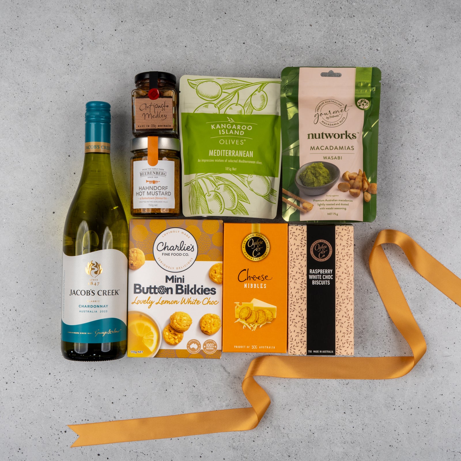 The Hamper Boutique Co's Gourmet Delights Hamper sits on a gray surface, filled with white wine, mustard, marmalade, olives, wasabi macadamias, lemon white choc cookies, cheese nibbles, raspberry choc biscuits, and finished with a yellow ribbon.