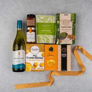 The Hamper Boutique Co's Gourmet Delights Hamper sits on a gray surface, filled with white wine, mustard, marmalade, olives, wasabi macadamias, lemon white choc cookies, cheese nibbles, raspberry choc biscuits, and finished with a yellow ribbon.