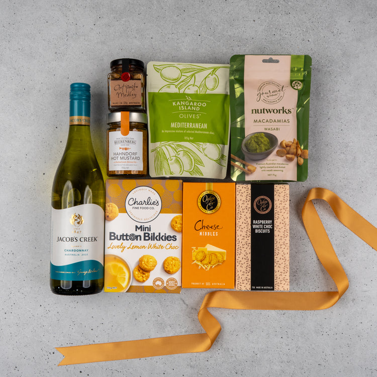 The Hamper Boutique Co's Gourmet Delights Hamper sits on a gray surface, filled with white wine, mustard, marmalade, olives, wasabi macadamias, lemon white choc cookies, cheese nibbles, raspberry choc biscuits, and finished with a yellow ribbon.
