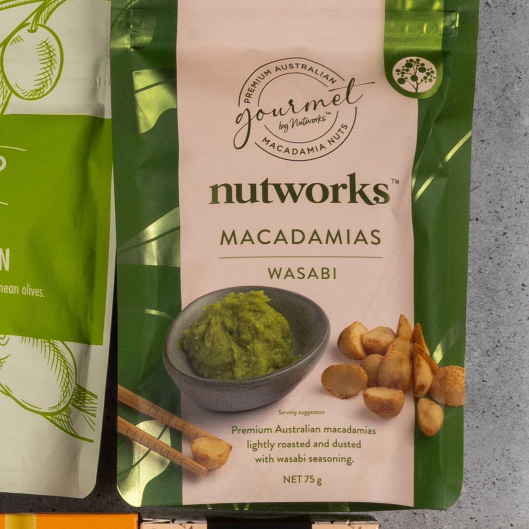 A green and cream package from The Hamper Boutique Co's Gourmet Delights Hamper is shown, featuring images of macadamia nuts and a bowl of green wasabi paste on the front. The package reads Premium Australian macadamias.