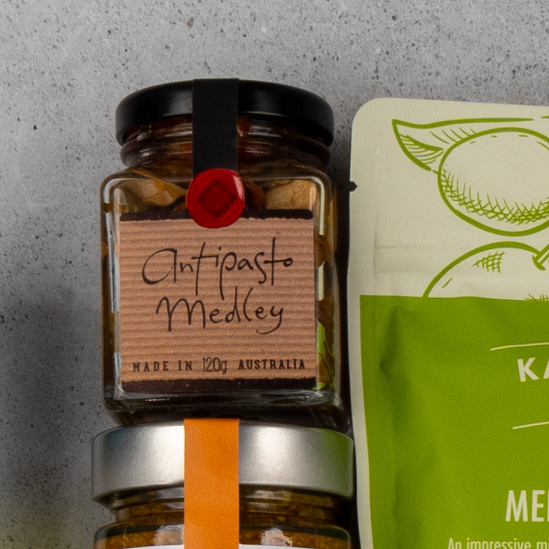 A jar labeled Gourmet Delights Hamper from The Hamper Boutique Co with a black lid and red seal sits beside a green and white food package featuring an illustrated animal head.
