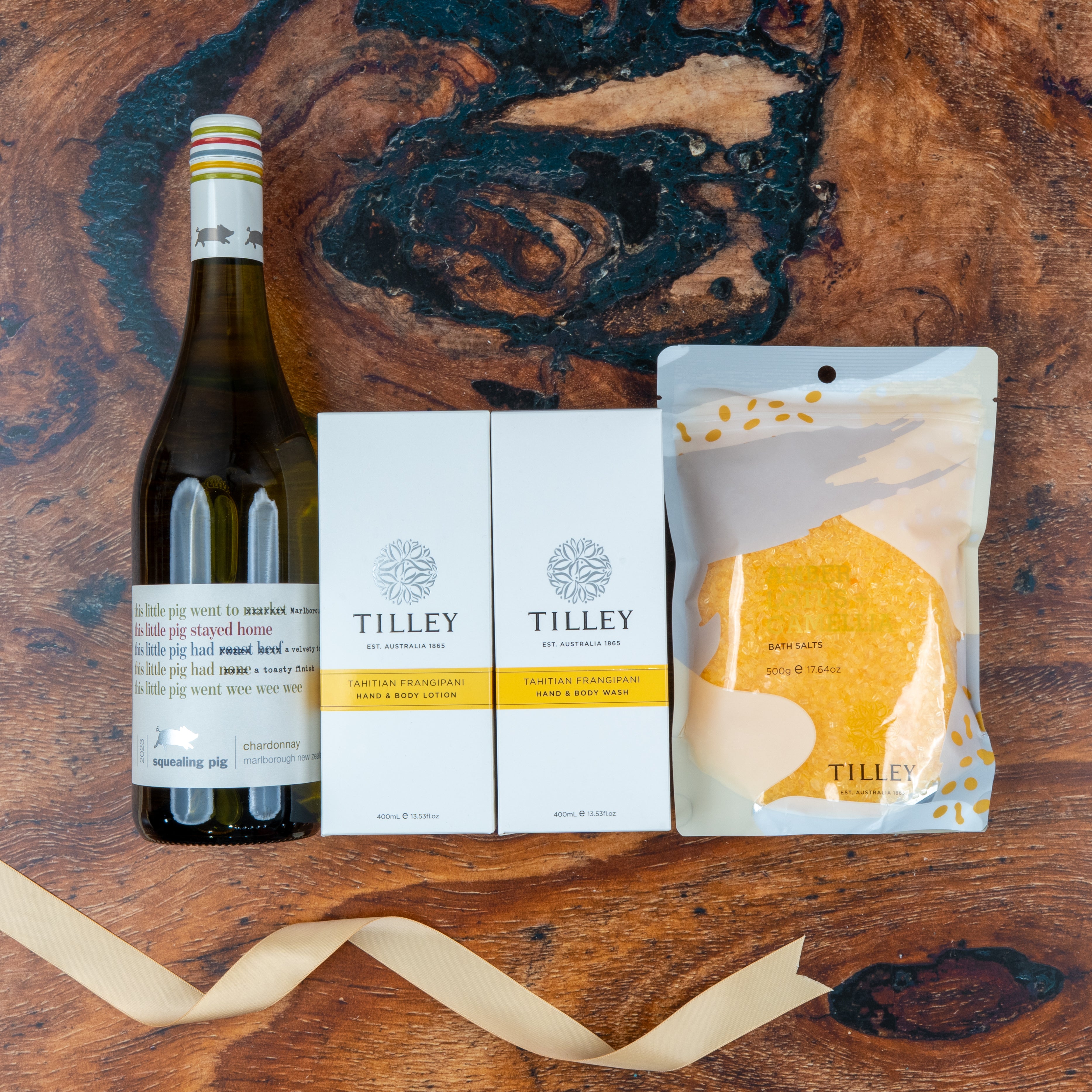 Chardonnay and Self-Care Essentials by The Hamper Boutique Co features a bottle of sparkling wine, two Tilley soap bars, and yellow bath salts, all beautifully arranged on a wooden surface with a gold ribbon in front.