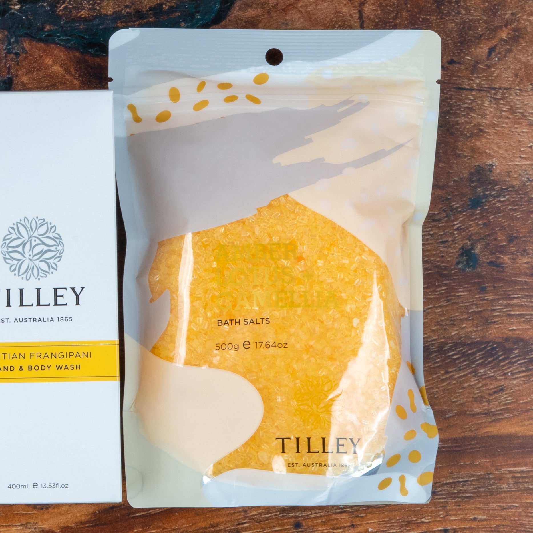 A clear resealable pouch labeled The Hamper Boutique Co Chardonnay and Self-Care Essentials with yellow bath salts sits on a wooden surface next to a white soap box.