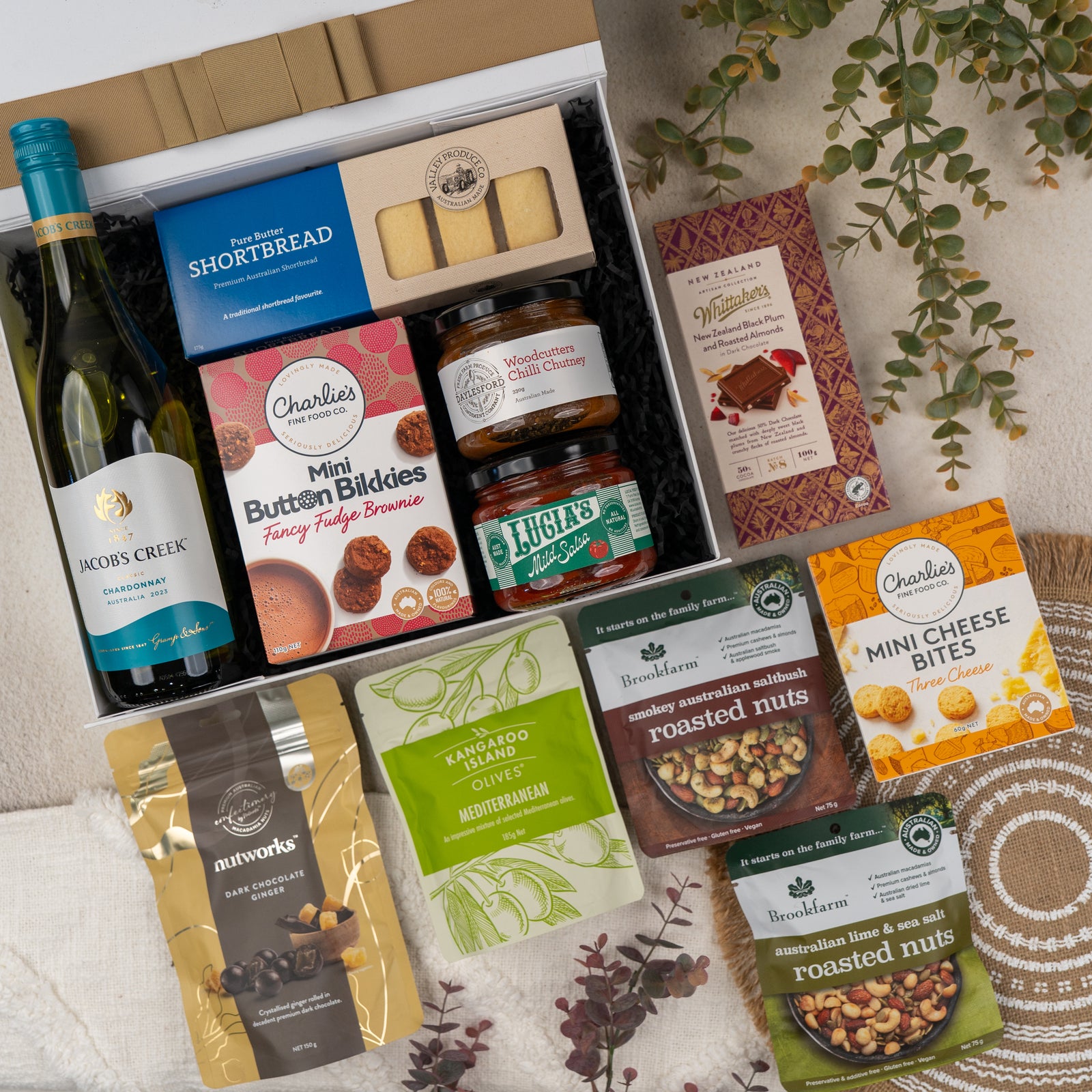 The Hamper Boutique Co’s Gourmet Indulgence Hamper includes Jacobs Creek Chardonnay, assorted snacks like shortbread, olives, nuts, cheese bites, fudge brownies, and chutney in a stylish box with decorative greenery.