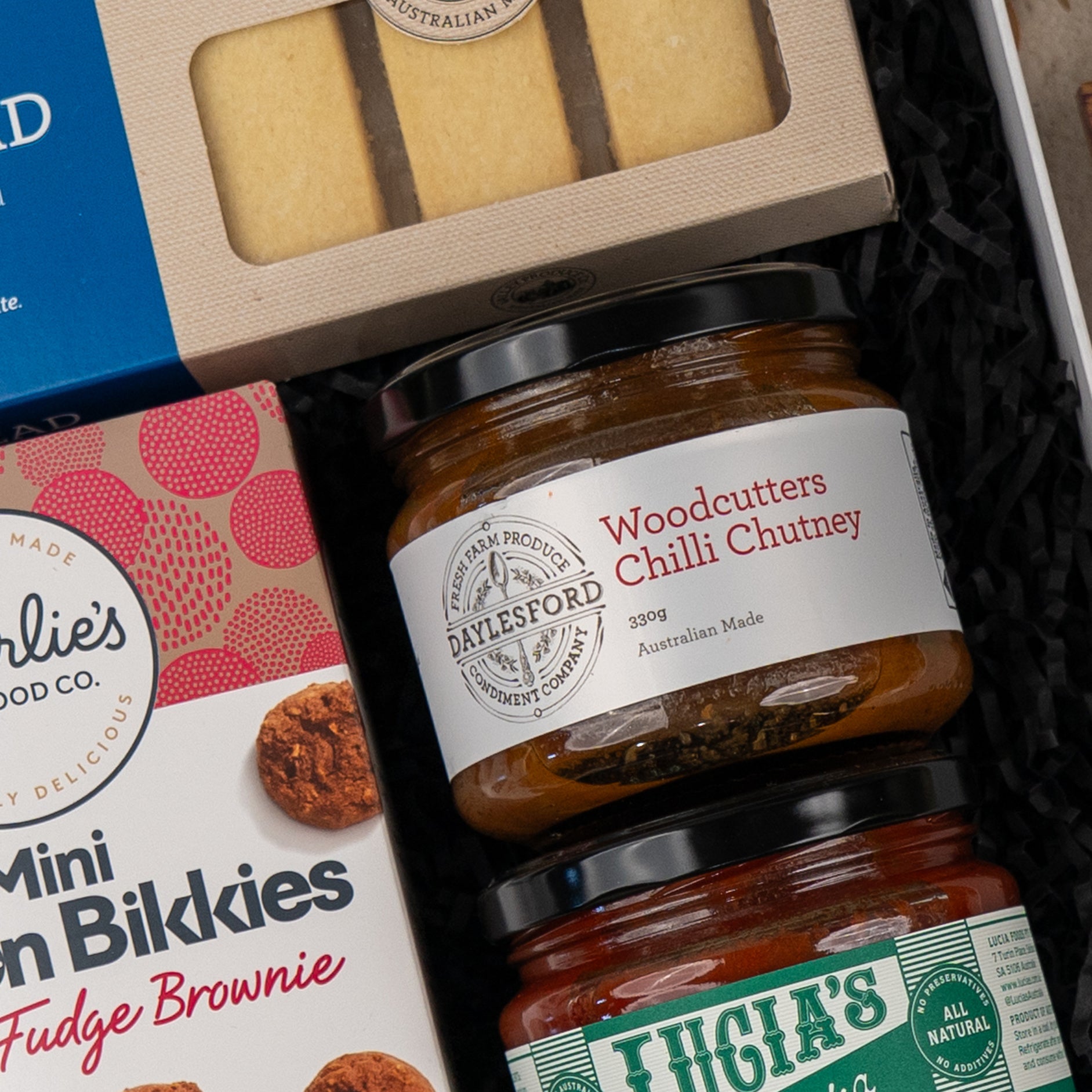 Close-up of The Hamper Boutique Co's Gourmet Indulgence Hamper, featuring Woodcutters Chilli Chutney jars, shortbread biscuits, and mini fudge brownies nestled in black shredded paper.
