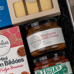 Close-up of The Hamper Boutique Co's Gourmet Indulgence Hamper, featuring Woodcutters Chilli Chutney jars, shortbread biscuits, and mini fudge brownies nestled in black shredded paper.