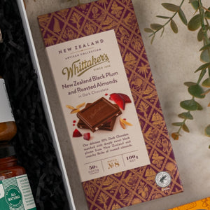 The Hamper Boutique Co's Gourmet Indulgence Hamper features a Whittakers New Zealand Black Plum and Roasted Almonds dark chocolate bar in elegant purple and gold packaging, beautifully presented in a gift box alongside a plant and assorted treats.
