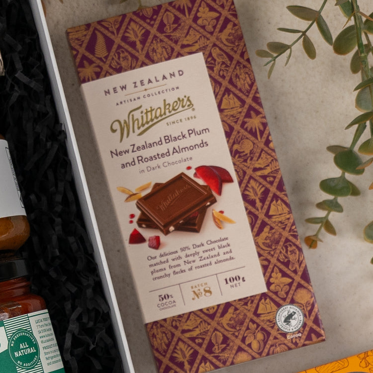 The Hamper Boutique Co's Gourmet Indulgence Hamper features a Whittakers New Zealand Black Plum and Roasted Almonds dark chocolate bar in elegant purple and gold packaging, beautifully presented in a gift box alongside a plant and assorted treats.