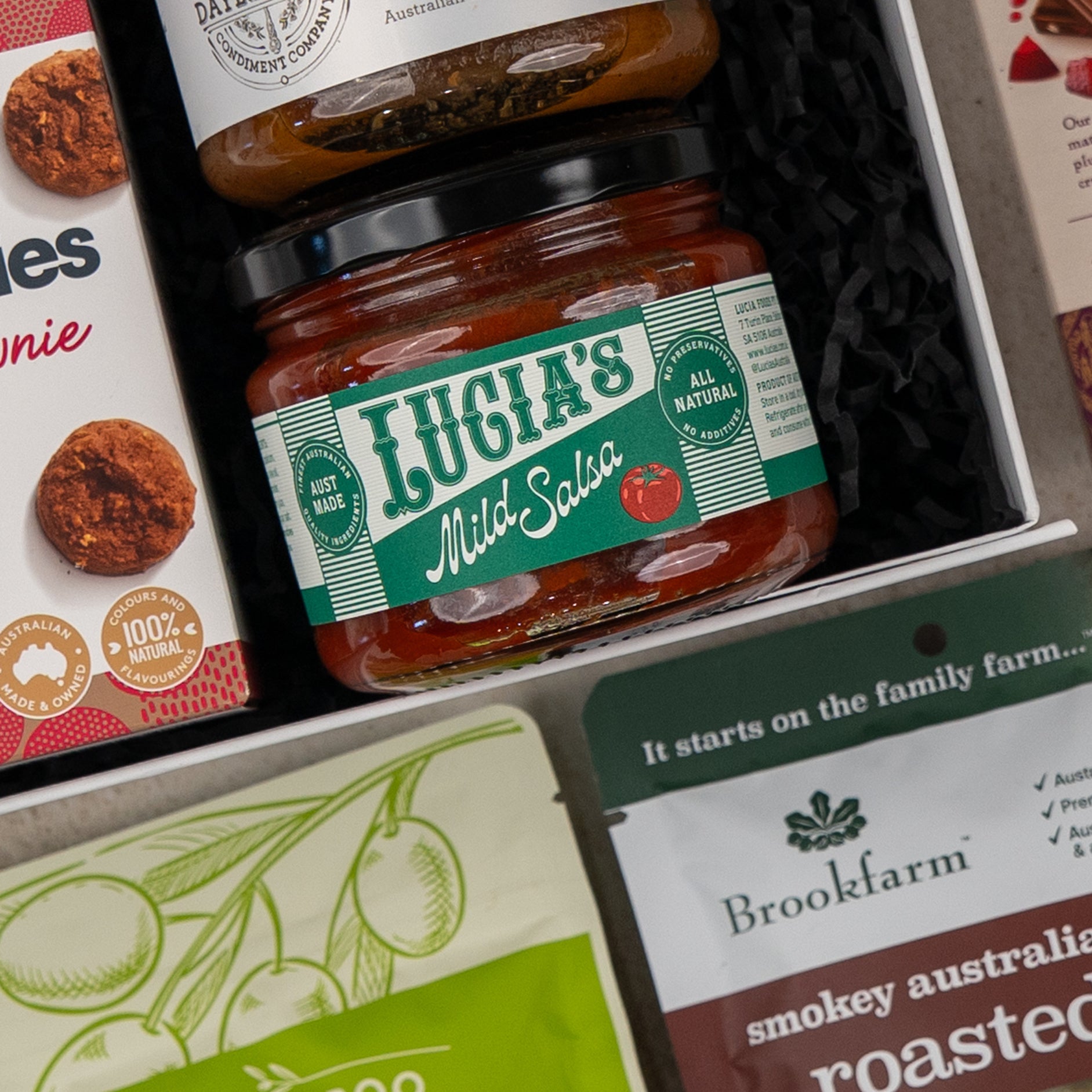 A close-up of the Gourmet Indulgence Hamper by The Hamper Boutique Co, featuring Lucias Mild Salsa, chutney, Brookfarm roasted nuts, and Byron Bay Cookies Triple Choc Fudge Brownie arranged on black shredded paper.