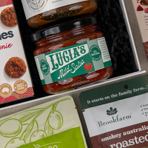 A close-up of the Gourmet Indulgence Hamper by The Hamper Boutique Co, featuring Lucias Mild Salsa, chutney, Brookfarm roasted nuts, and Byron Bay Cookies Triple Choc Fudge Brownie arranged on black shredded paper.