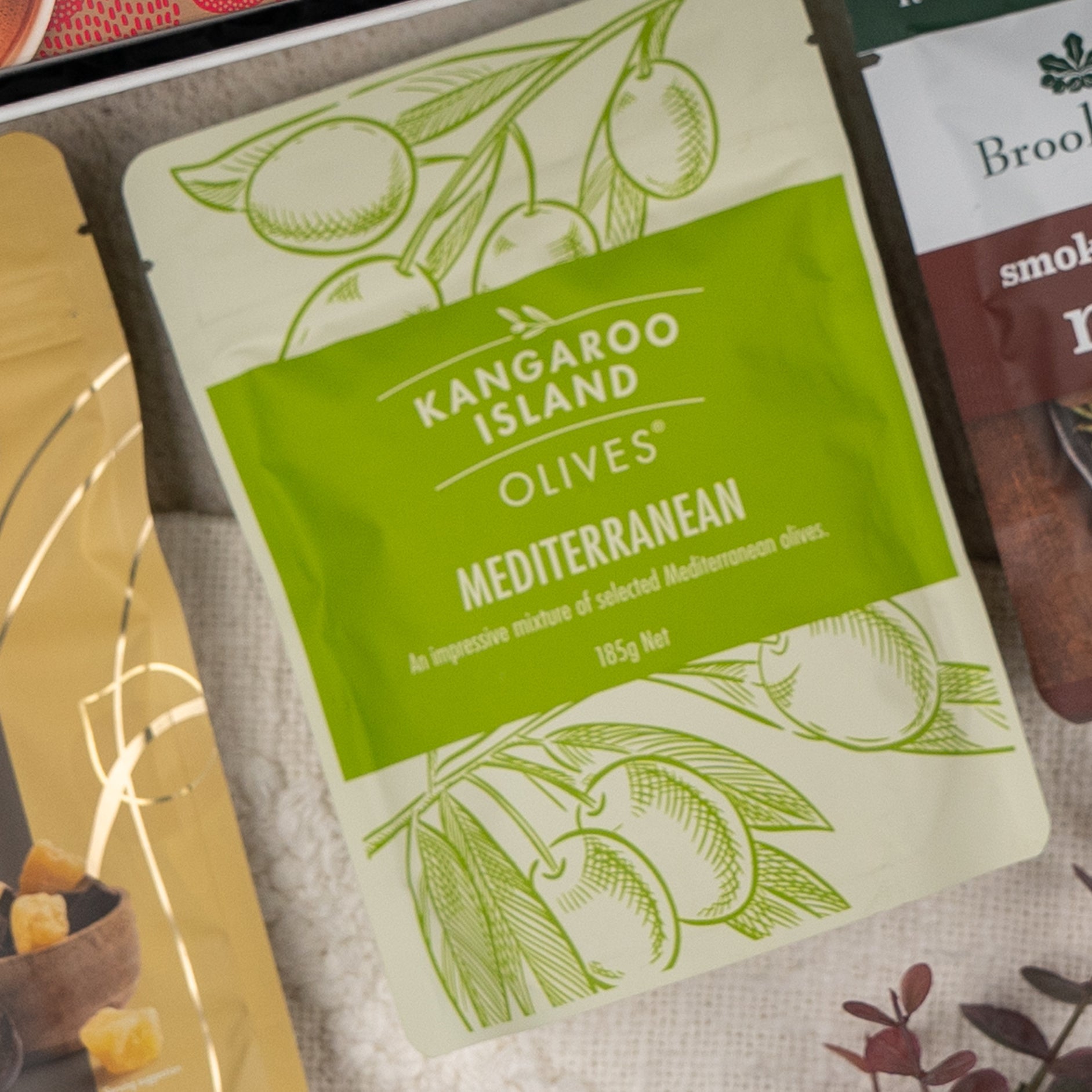 A Gourmet Indulgence Hamper from The Hamper Boutique Co is placed among other packaged food items on a light-colored surface.
