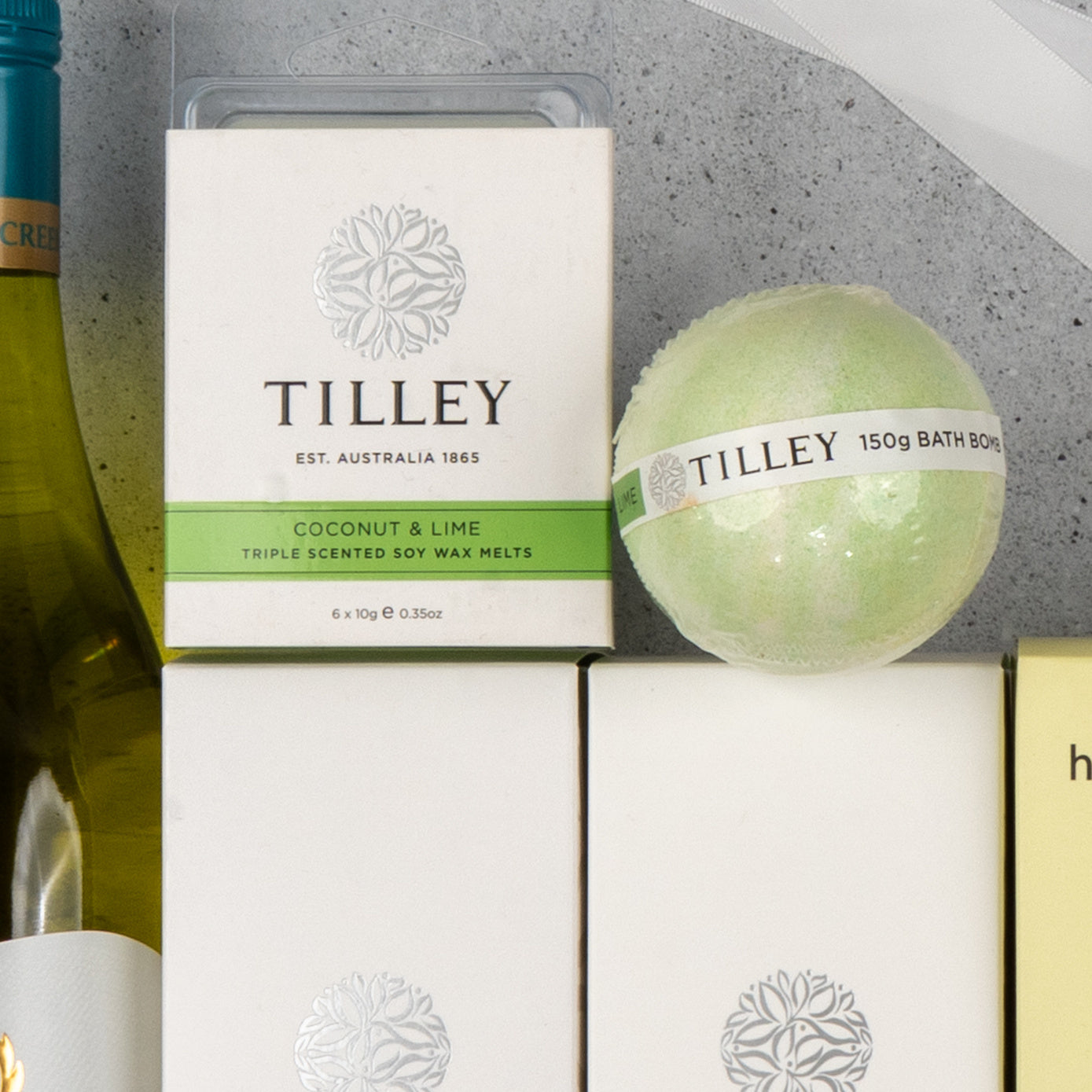 A close-up of The Hamper Boutique Co's "White Wine and Self-Care" set, featuring Coconut & Lime wax melts and a green bath bomb, displayed with a wine bottle on a light gray surface.