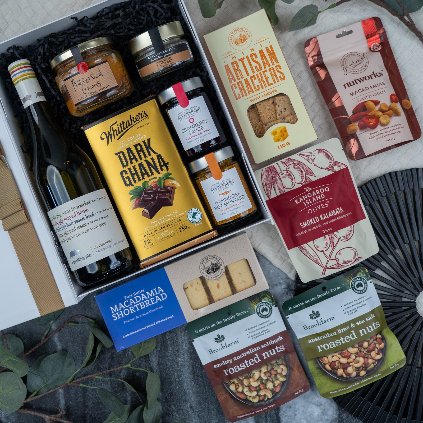 The Hamper Boutique Co's Gourmet Experience Hamper features wine, dark chocolate, crackers, jams, olives, macadamia shortbread, roasted nuts, and macadamia caramel clusters arranged elegantly with green leaves.
