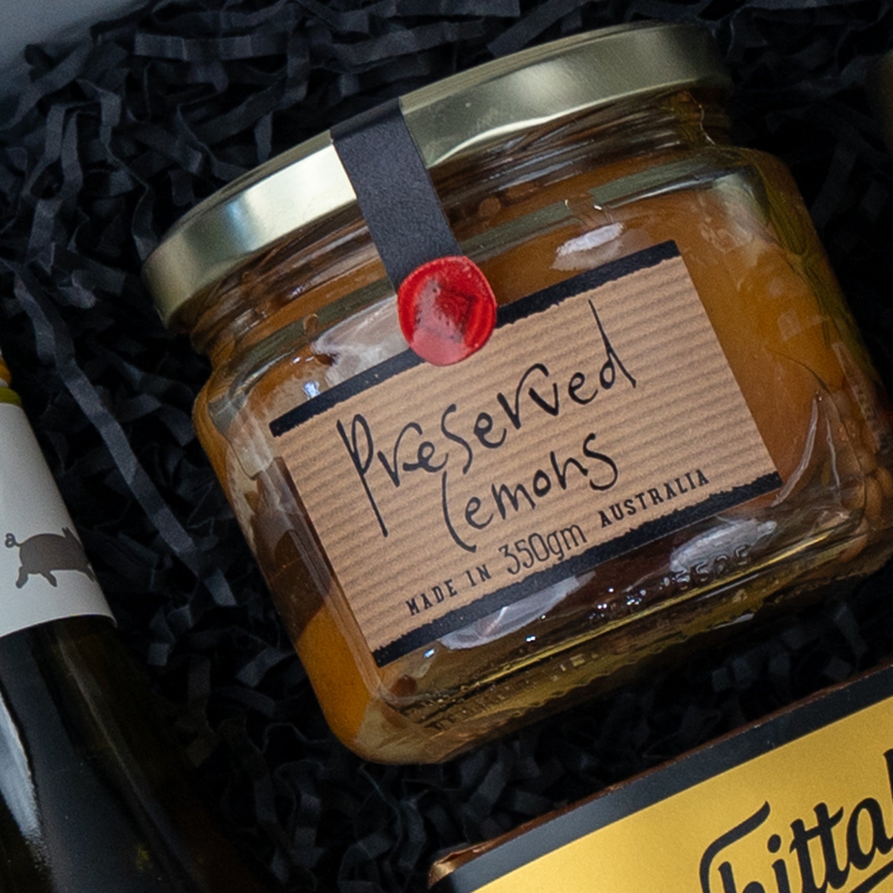 A glass jar labeled Preserved Lemons with a gold lid, weighing 350g and made in Australia, from The Hamper Boutique Co’s Gourmet Experience Hamper, sits on black crinkle paper. The label features handwritten text.