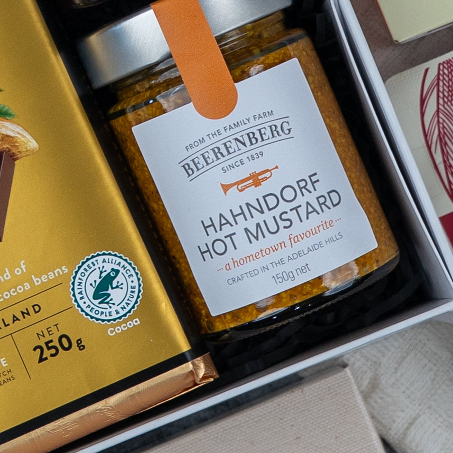 The Hamper Boutique Co's Gourmet Experience Hamper features a gold-wrapped chocolate bar and a jar of Beerenberg Hahndorf Hot Mustard with a white label and orange seal, both elegantly arranged in the box.