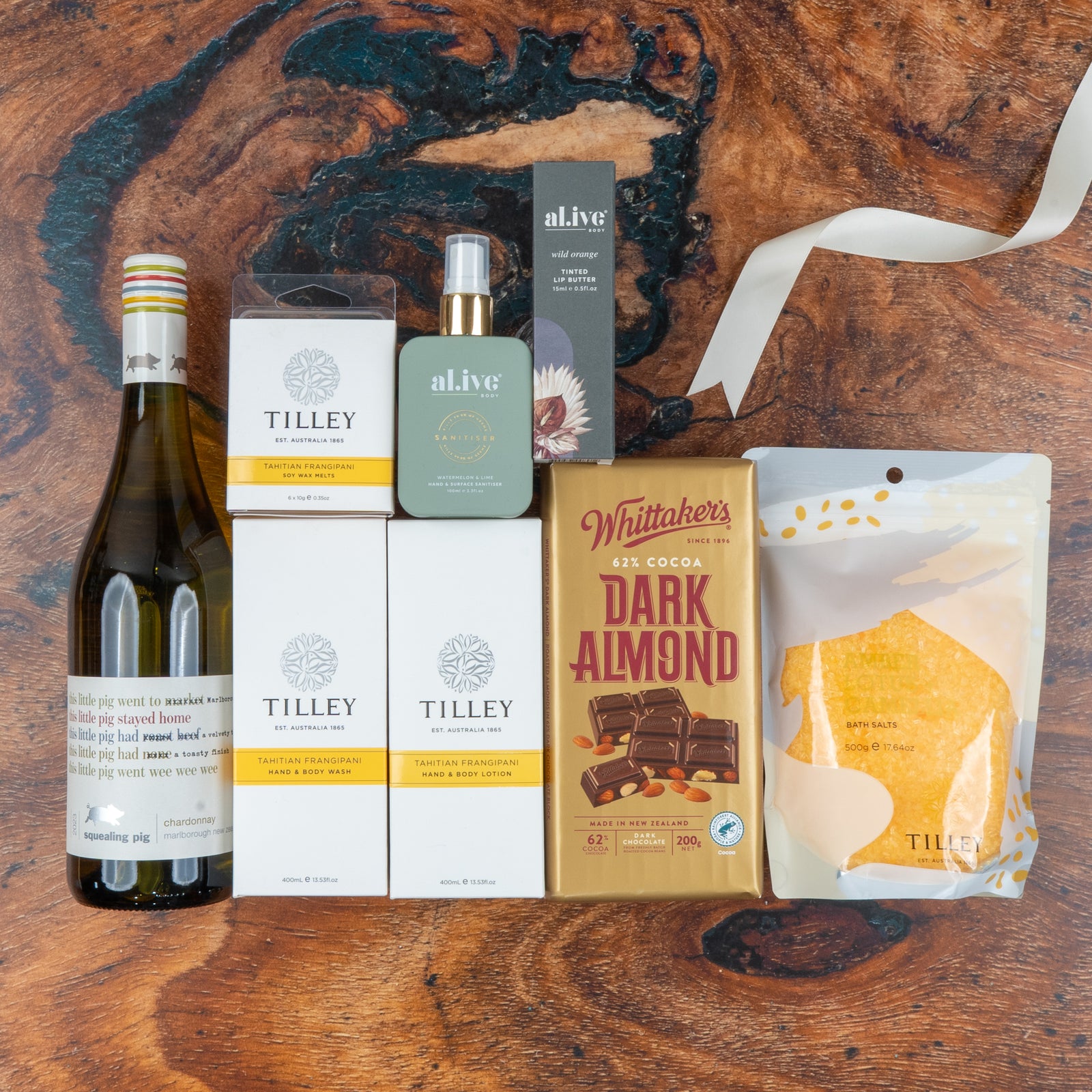 A flat lay of the Chardonnay and Self-Care Hamper by The Hamper Boutique Co, featuring white wine, four Tilley product boxes, Whittakers dark almond chocolate, bath salts, and a.live room spray on a wooden surface.
