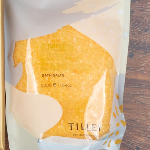 A clear plastic pouch of yellow bath salts from The Hamper Boutique Co’s “Chardonnay and Self-Care Hamper” sits on a wooden surface.