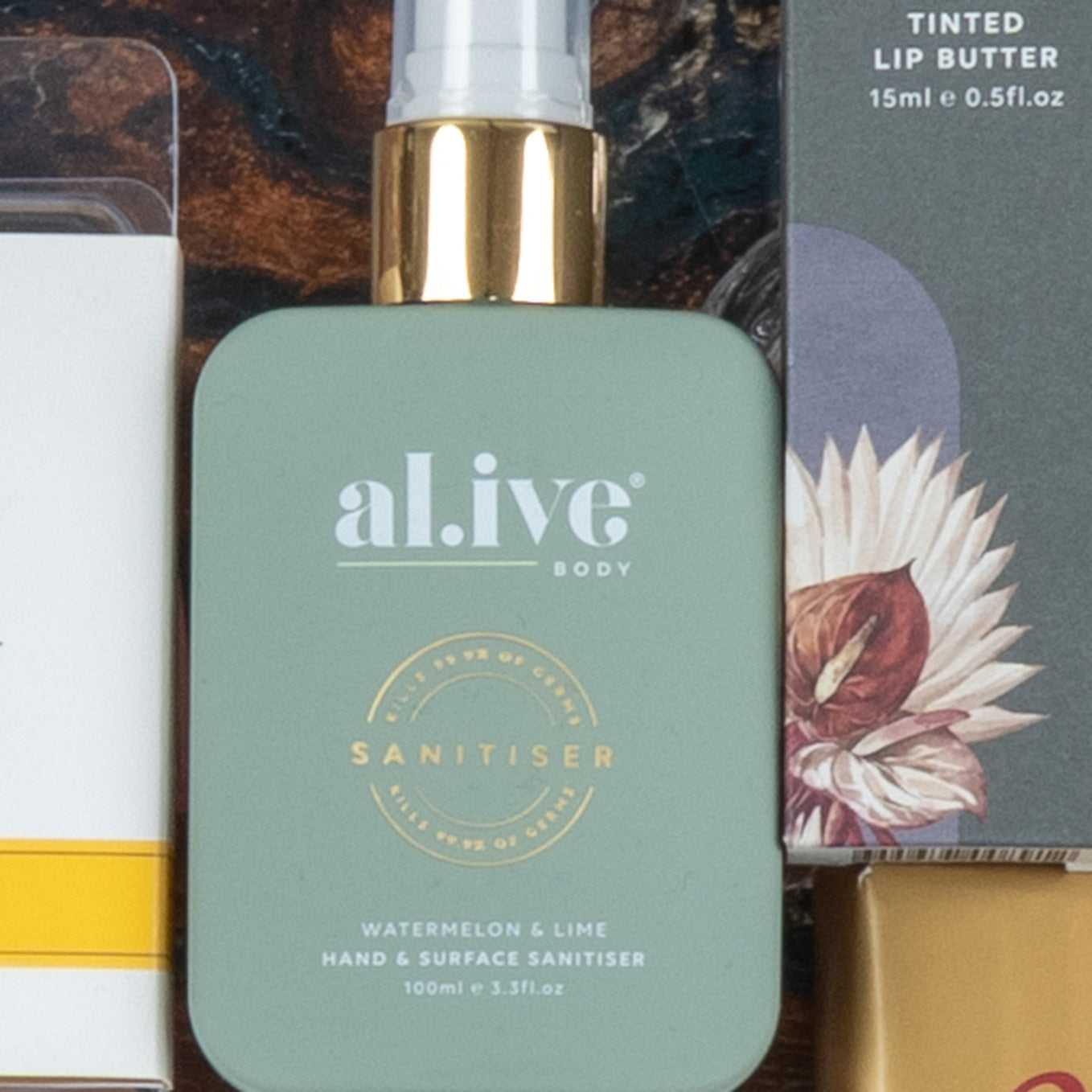 A close-up of The Hamper Boutique Co's Chardonnay and Self-Care Hamper, showing a green al.ive body Sanitiser with gold spray top (watermelon & lime scent) beside a box of tinted lip butter decorated with a floral illustration.