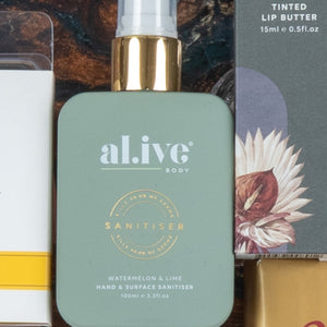 A close-up of The Hamper Boutique Co's Chardonnay and Self-Care Hamper, showing a green al.ive body Sanitiser with gold spray top (watermelon & lime scent) beside a box of tinted lip butter decorated with a floral illustration.