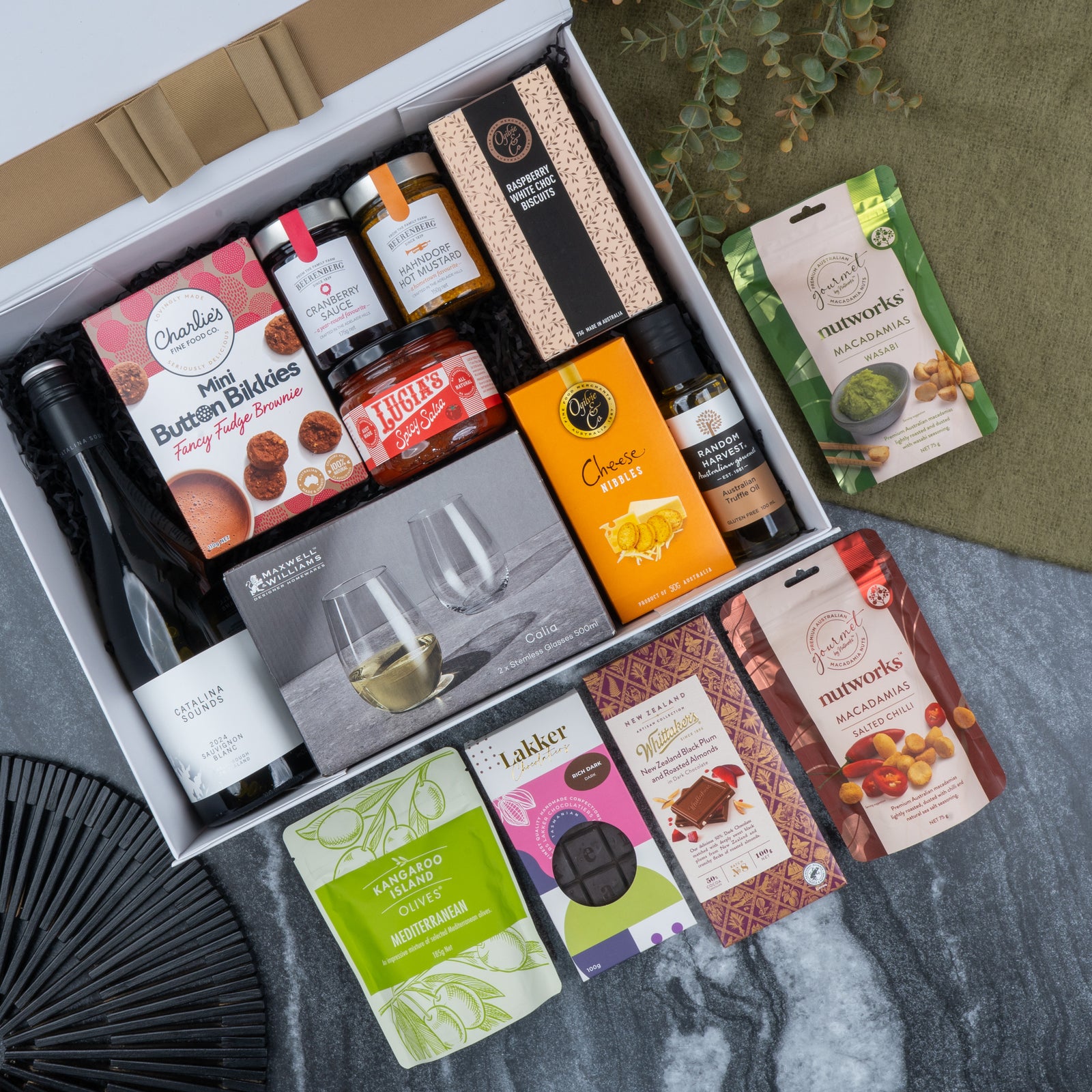 The Hamper Boutique Co's Epicurean Delights Hamper features wine, chocolates, cookies, nuts, crackers, preserves, cheese crisps, and two clear wine glasses—beautifully arranged on a marble and fabric surface.