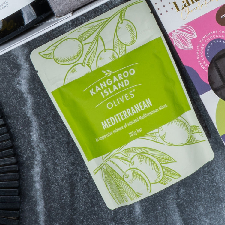 A green and white pouch from The Hamper Boutique Co labeled Epicurean Delights Hamper lies on a dark marble surface, featuring olive branch illustrations and containing 185g of mixed Mediterranean olives.