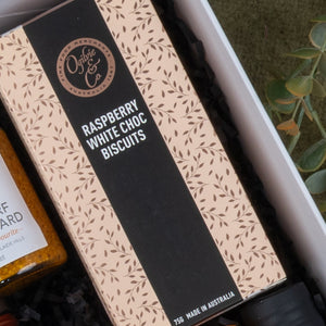 An Epicurean Delights Hamper by The Hamper Boutique Co features Raspberry White Choc Biscuits in a beige and black leaf design box, nestled with black shredded paper. A jar can be partially seen to the left.
