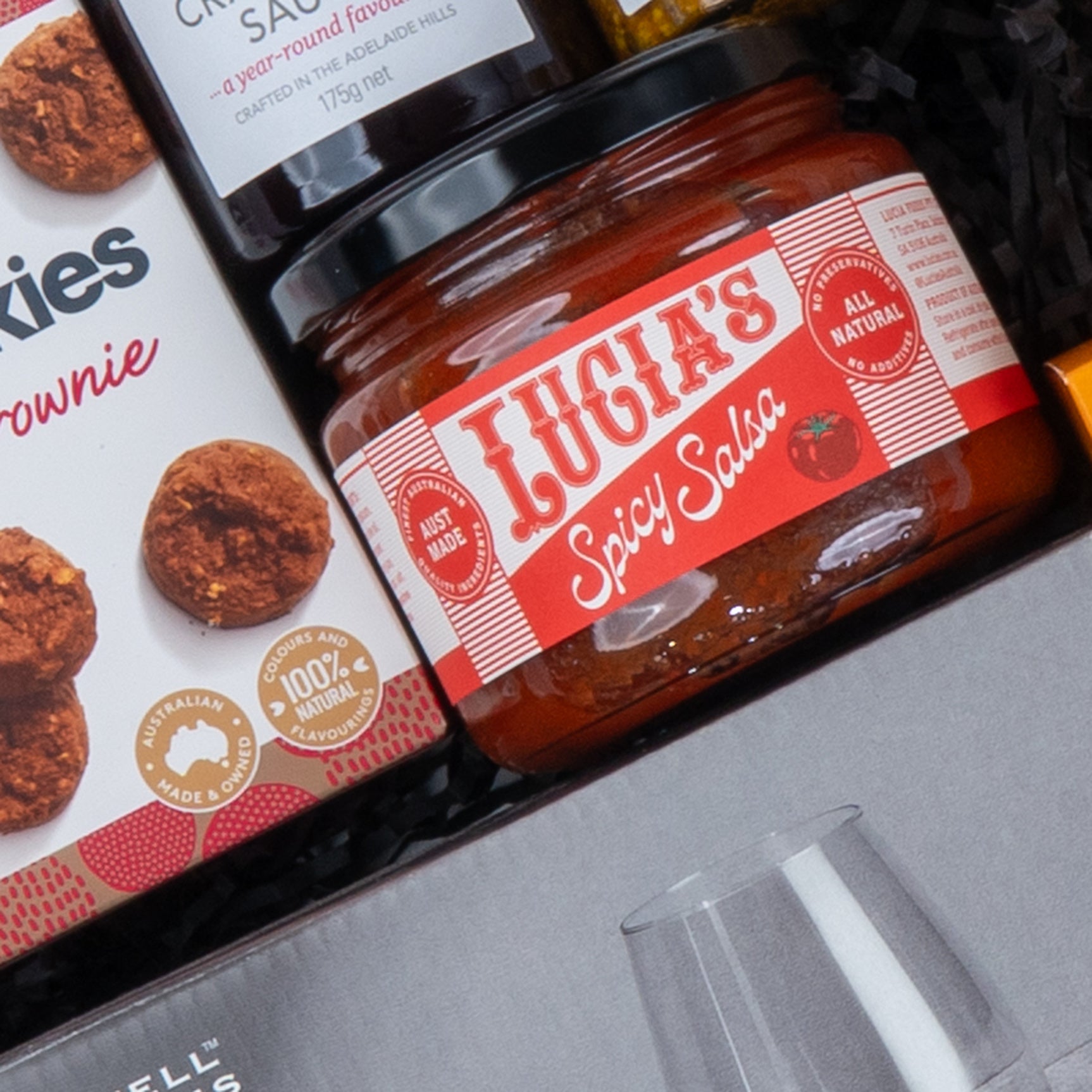 Close-up of the Epicurean Delights Hamper by The Hamper Boutique Co, featuring a jar of spicy salsa, snack brownies, and chutney attractively arranged on shredded black paper.
