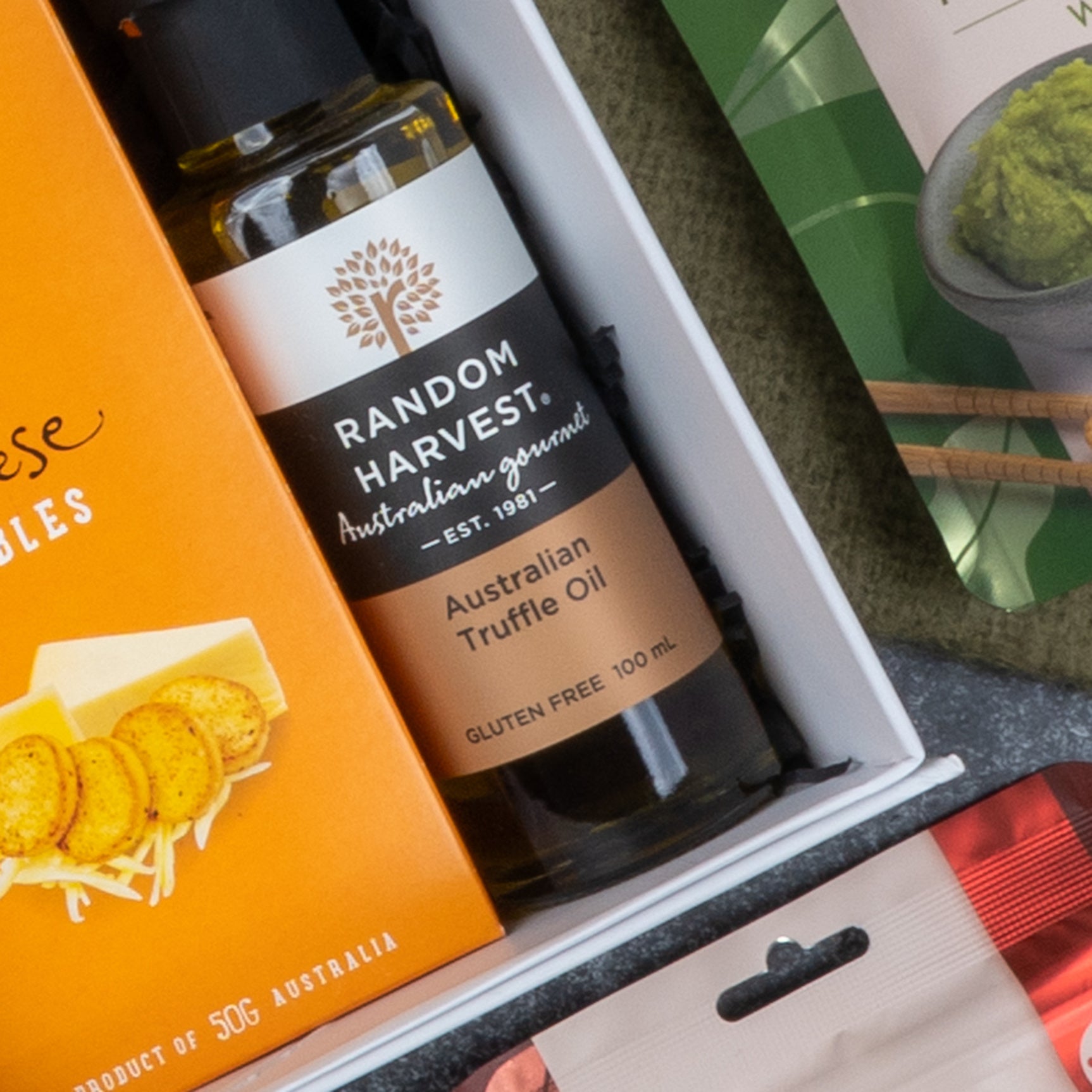 A close-up of The Hamper Boutique Co’s Epicurean Delights Hamper, featuring Random Harvest Australian Truffle Oil, cheese nibbles in an orange box, and part of a green food package.