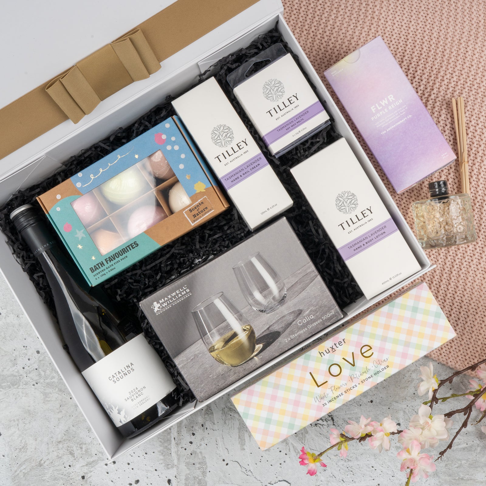The Chardonnay and Relaxation Gift Hamper by The Hamper Boutique Co includes bath bombs, scented candles, Tilley soaps, a small wine bottle, stemless wine glasses, reed diffuser, and a pastel hustle Love box with cherry blossom decor.