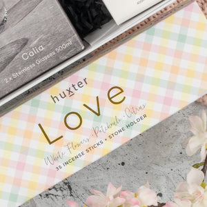 A close-up of The Hamper Boutique Co Chardonnay and Relaxation Gift Hamper, featuring incense sticks, stone holder, and stemless glasses. Soft pink flowers appear in the corner.