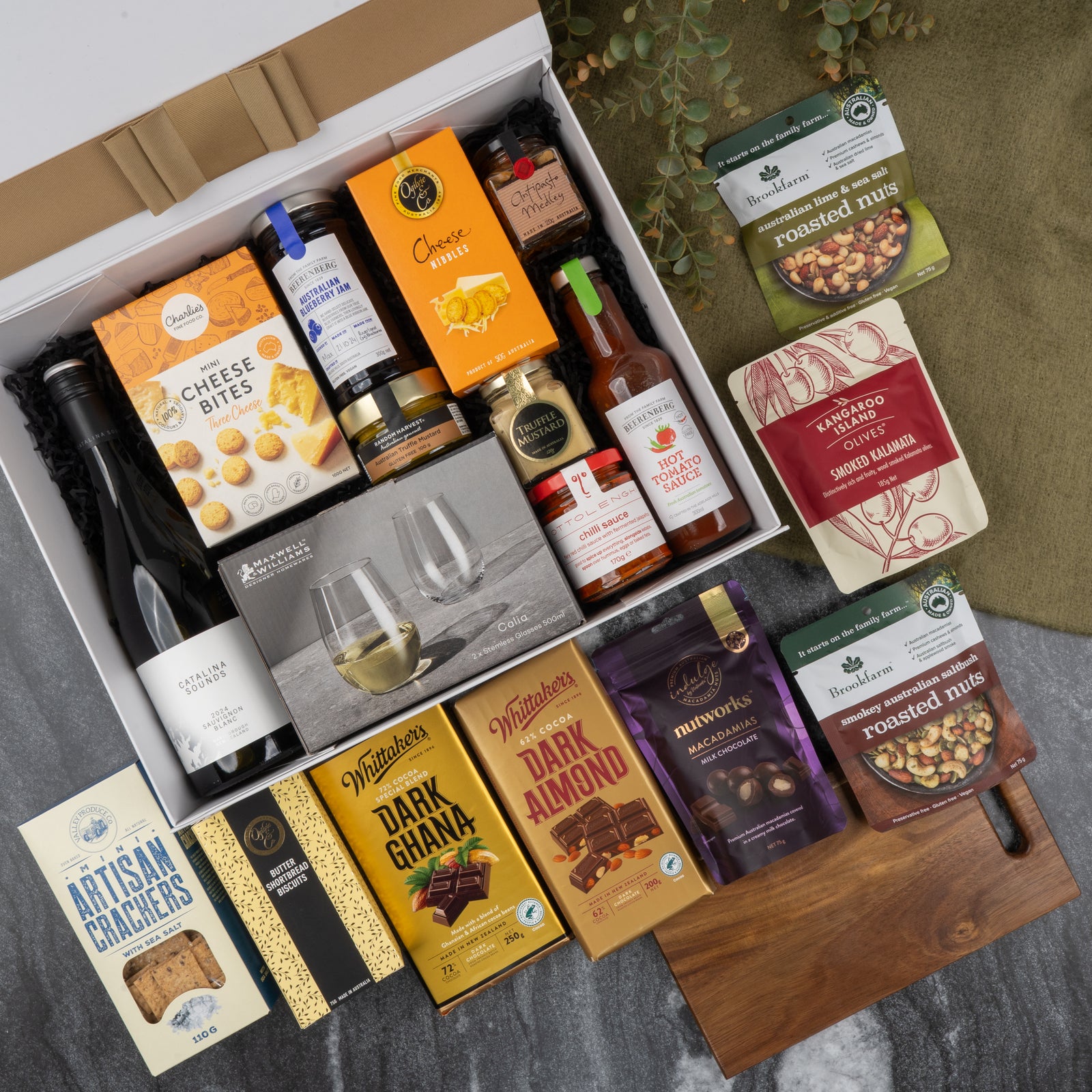 The Hamper Boutique Co's Gourmet Collection Gift Hamper includes wine, cheese bites, crackers, chocolates, mixed nuts, sauces, and drinkware in a white box with a wooden cutting board elegantly displayed on marble.