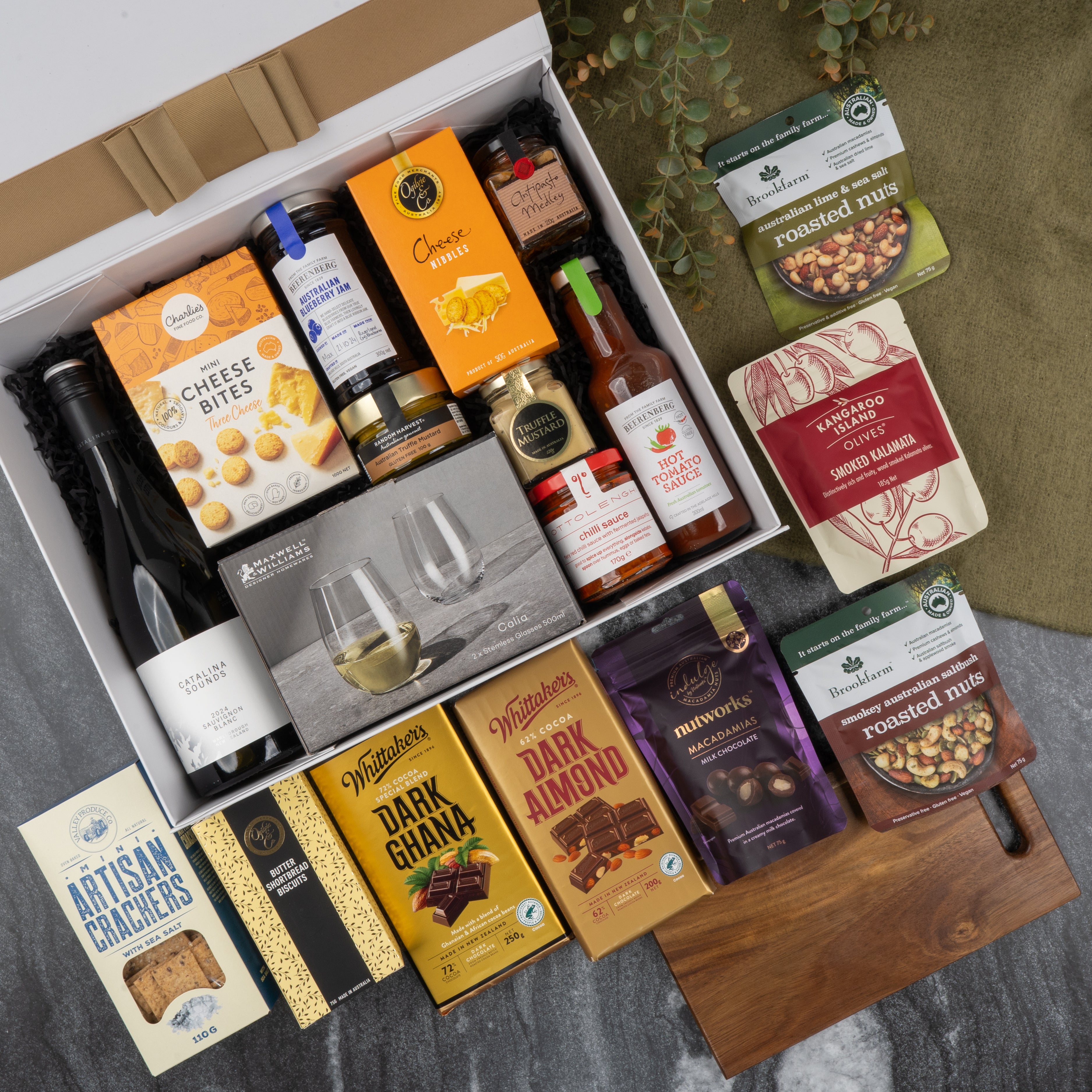 The Hamper Boutique Co's Gourmet Collection Gift Hamper includes wine, cheese bites, crackers, chocolates, mixed nuts, sauces, and drinkware in a white box with a wooden cutting board elegantly displayed on marble.