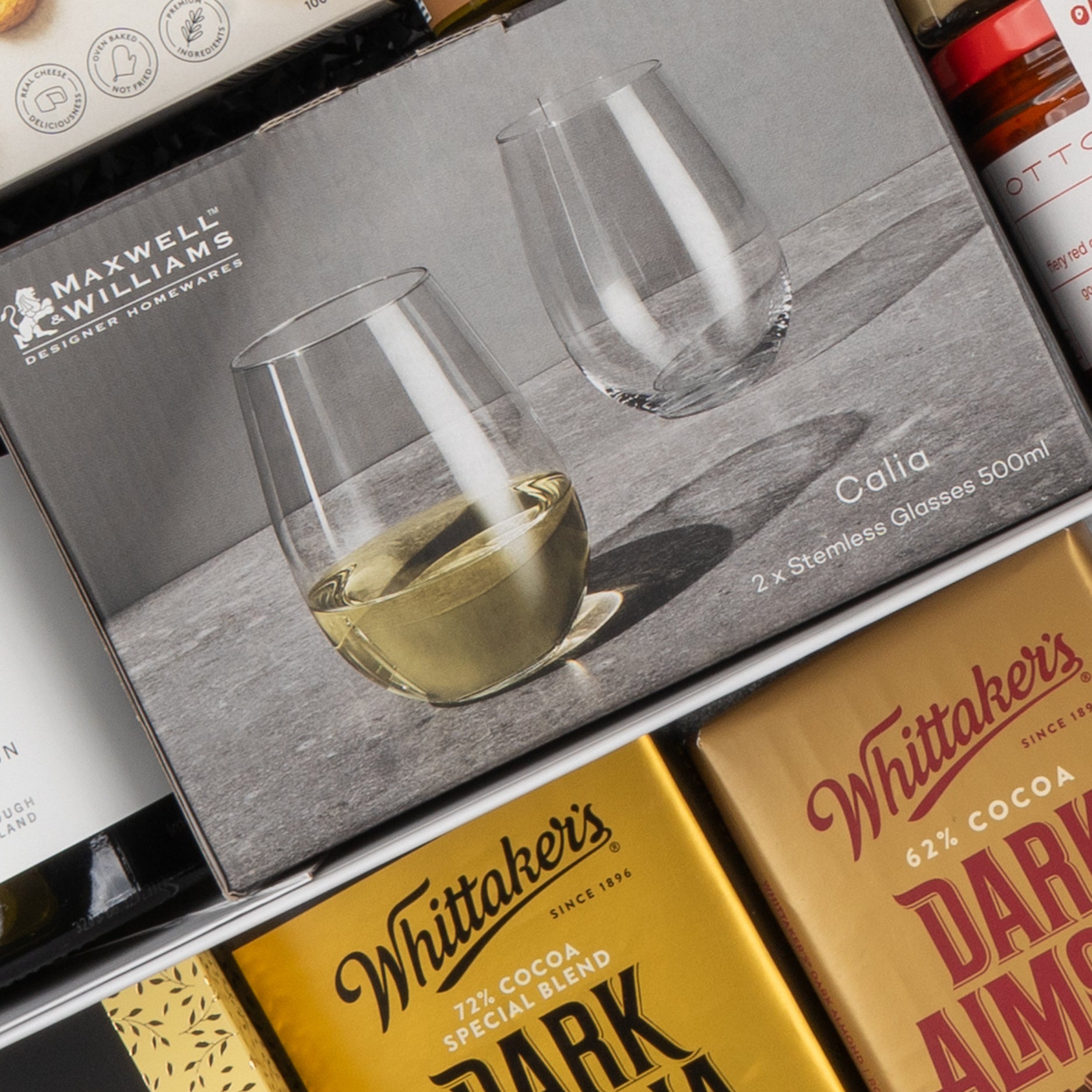 The Hamper Boutique Co’s Gourmet Collection Gift Hamper features a boxed set of two Maxwell & Williams Calla stemless glasses, one shown with white wine, displayed alongside Whittaker’s chocolate bars and assorted packaged gourmet treats.
