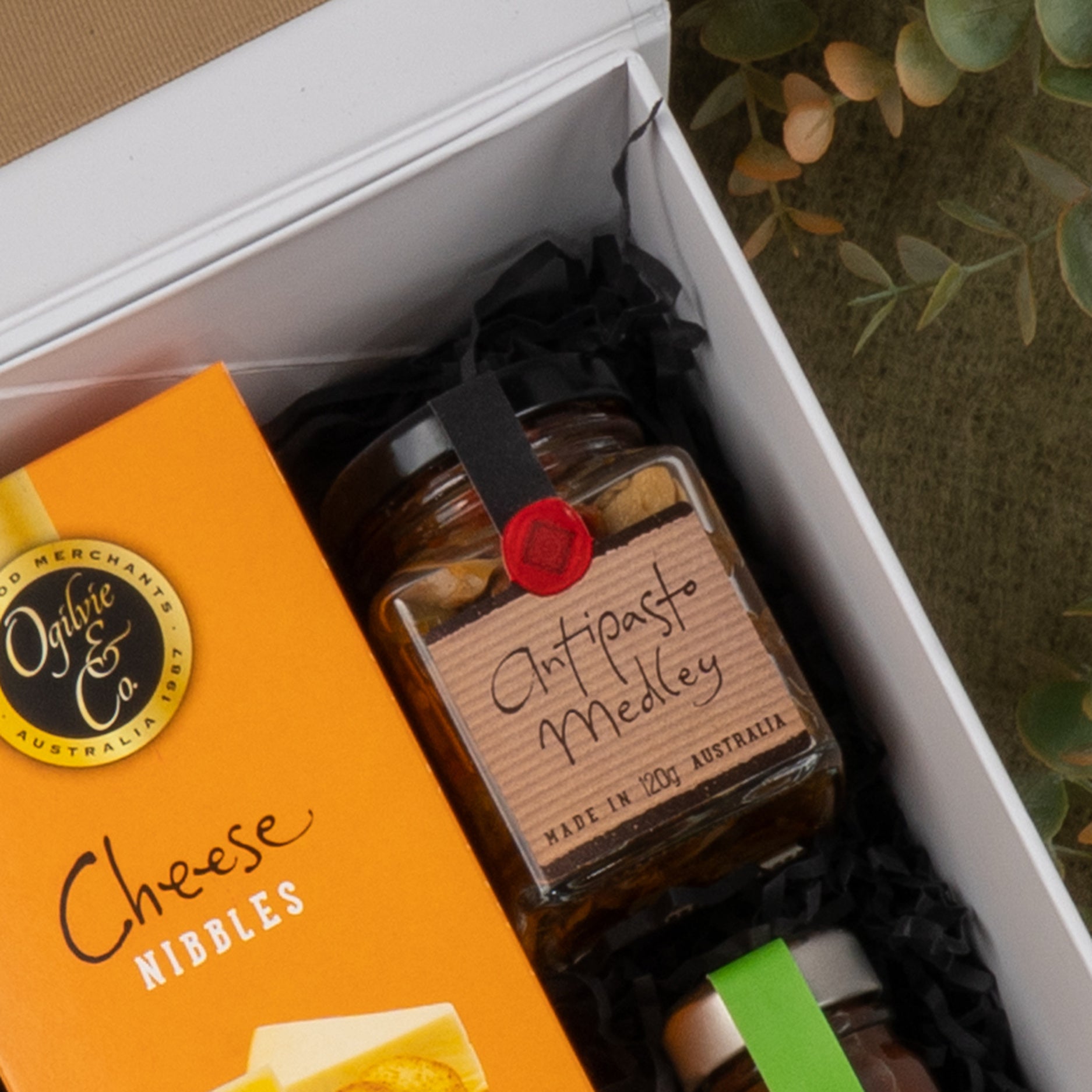 The Hamper Boutique Co's Gourmet Collection Gift Hamper features Antipasto Medley, cheese nibbles, and a small green-lidded jar, all arranged on black shredded paper beside green foliage.