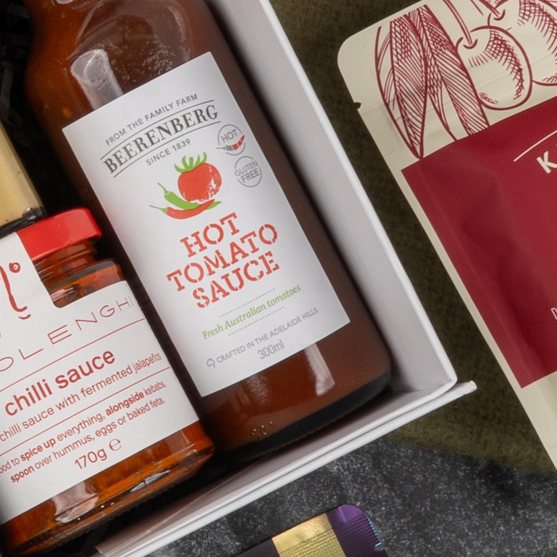 Close-up of The Hamper Boutique Co's Gourmet Collection Gift Hamper, featuring Beerenberg hot tomato sauce and a chili-labeled jar beside a red and cream package, all neatly arranged in the box.
