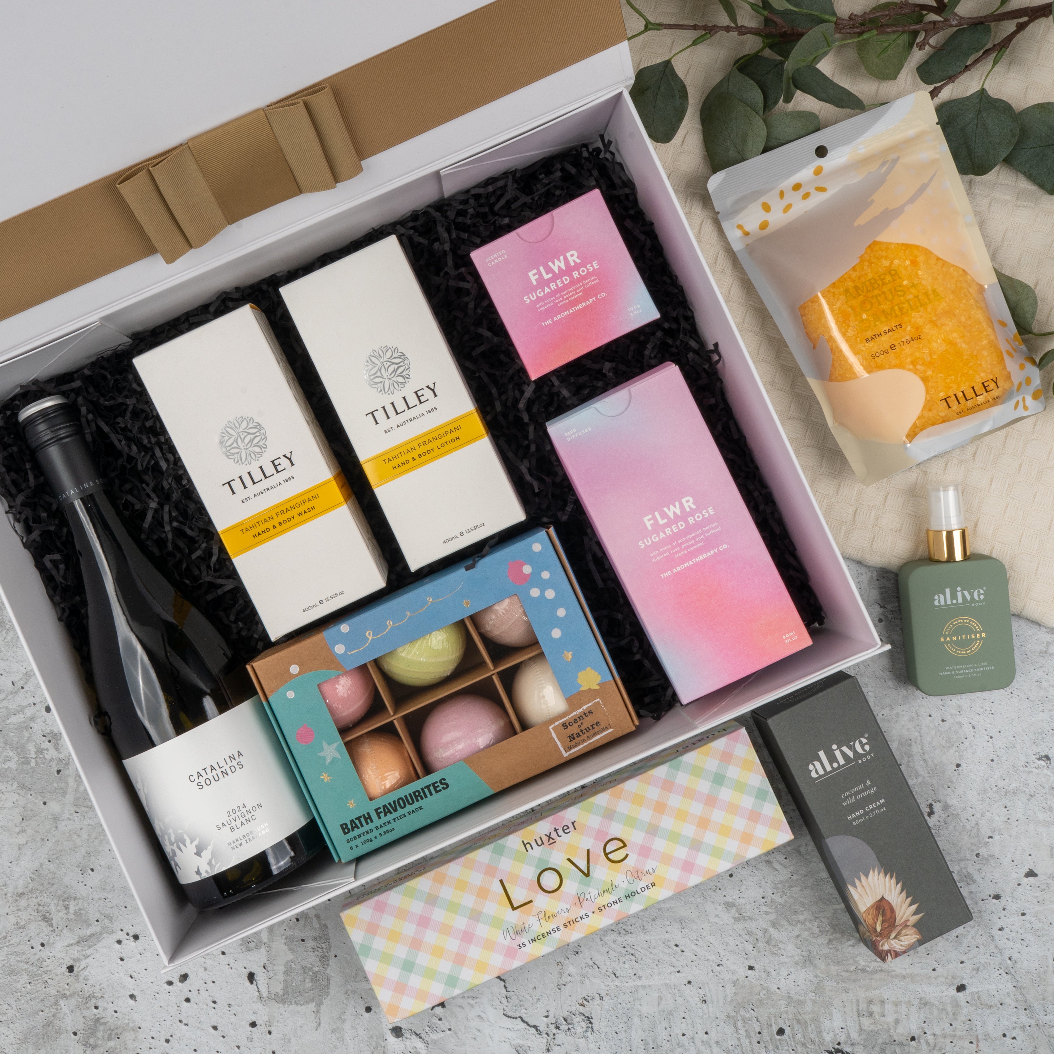 The Hamper Boutique Co's Ultimate Self-Care and Chardonnay Hamper features wine, Tilley soaps, FLWR bath and shower products, bath bombs, Alive skincare, and honeycomb, beautifully arranged on marble with greenery.