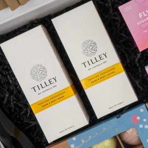 Two boxes from The Hamper Boutique Co’s Ultimate Self-Care and Chardonnay Hamper rest on black crinkle paper, with other colorful gift items partially visible nearby.