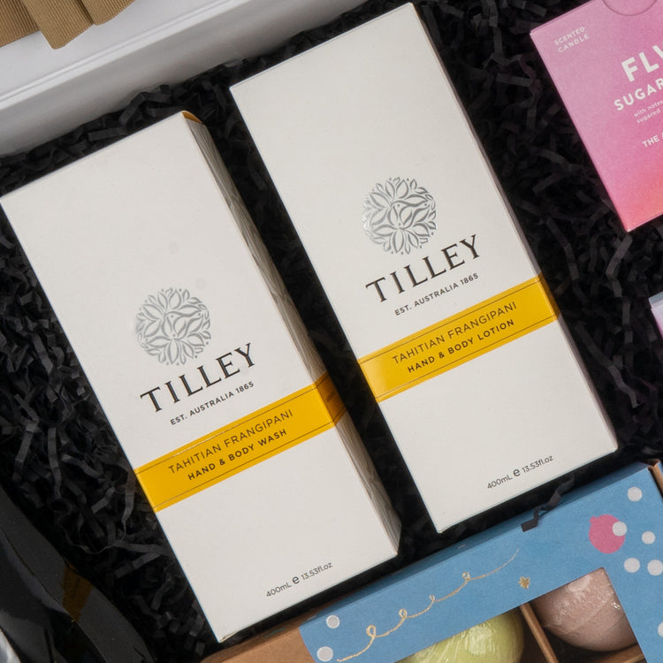 Two boxes from The Hamper Boutique Co’s Ultimate Self-Care and Chardonnay Hamper rest on black crinkle paper, with other colorful gift items partially visible nearby.