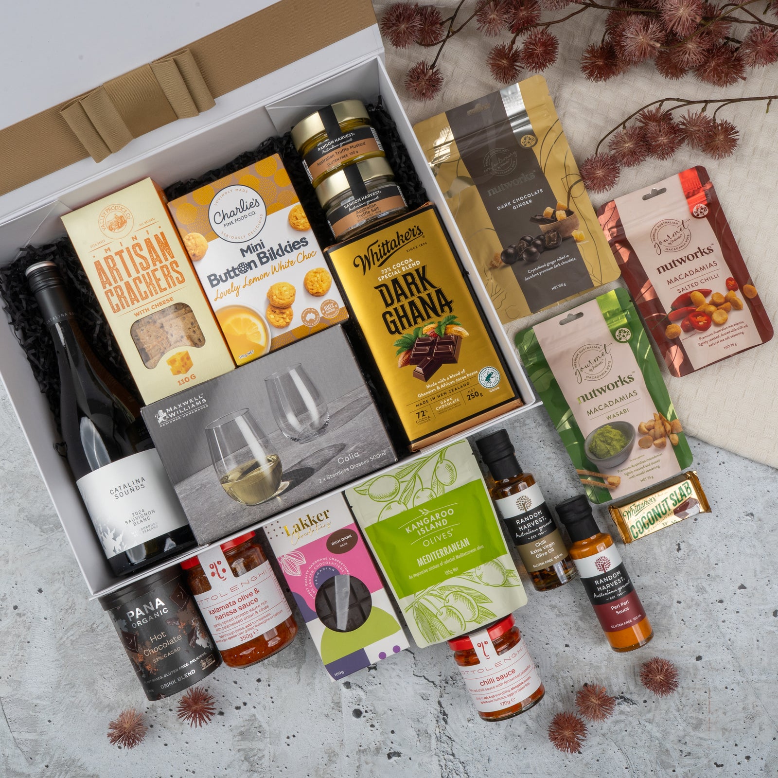 The Hamper Boutique Co’s Ultimate Gourmet Collection Hamper features wine, crackers, chocolates, mixed nuts, cookies, olive oil, mustard, sauces, and two wine glasses in a gift box adorned with dried flowers on a textured surface.