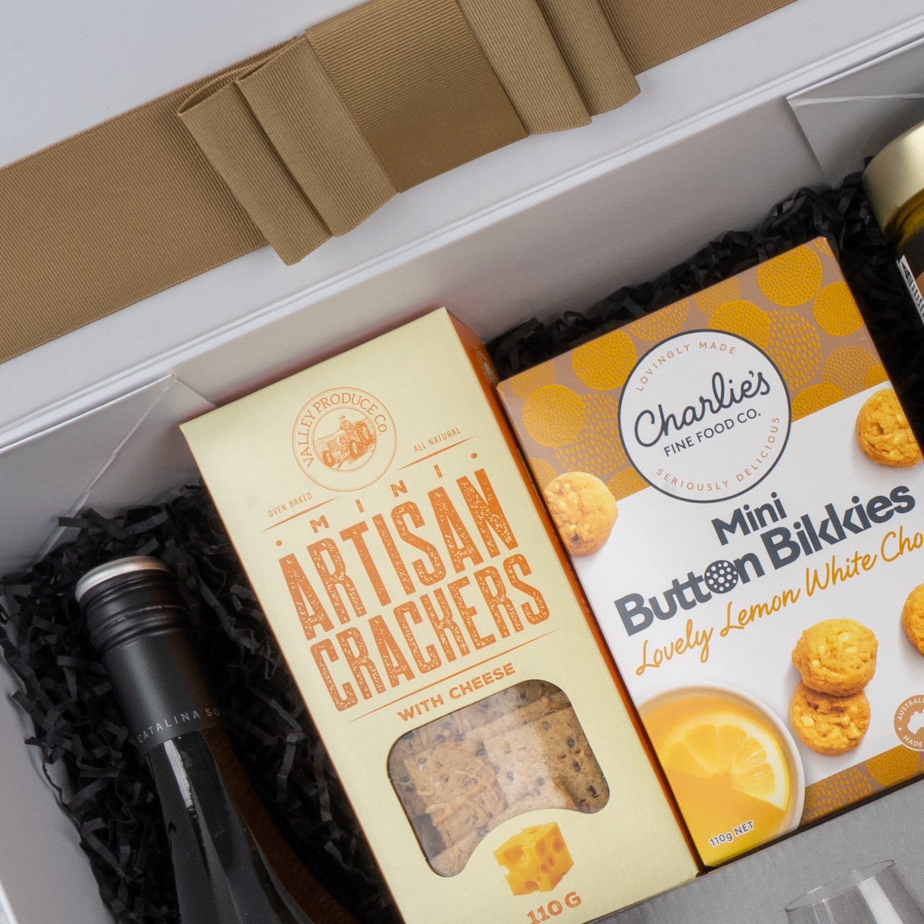 The Ultimate Gourmet Collection Hamper by The Hamper Boutique Co features wine, mini artisan cheese crackers, and lemon white chocolate button biscuits, beautifully arranged on black shredded paper with a beige ribbon on the lid.
