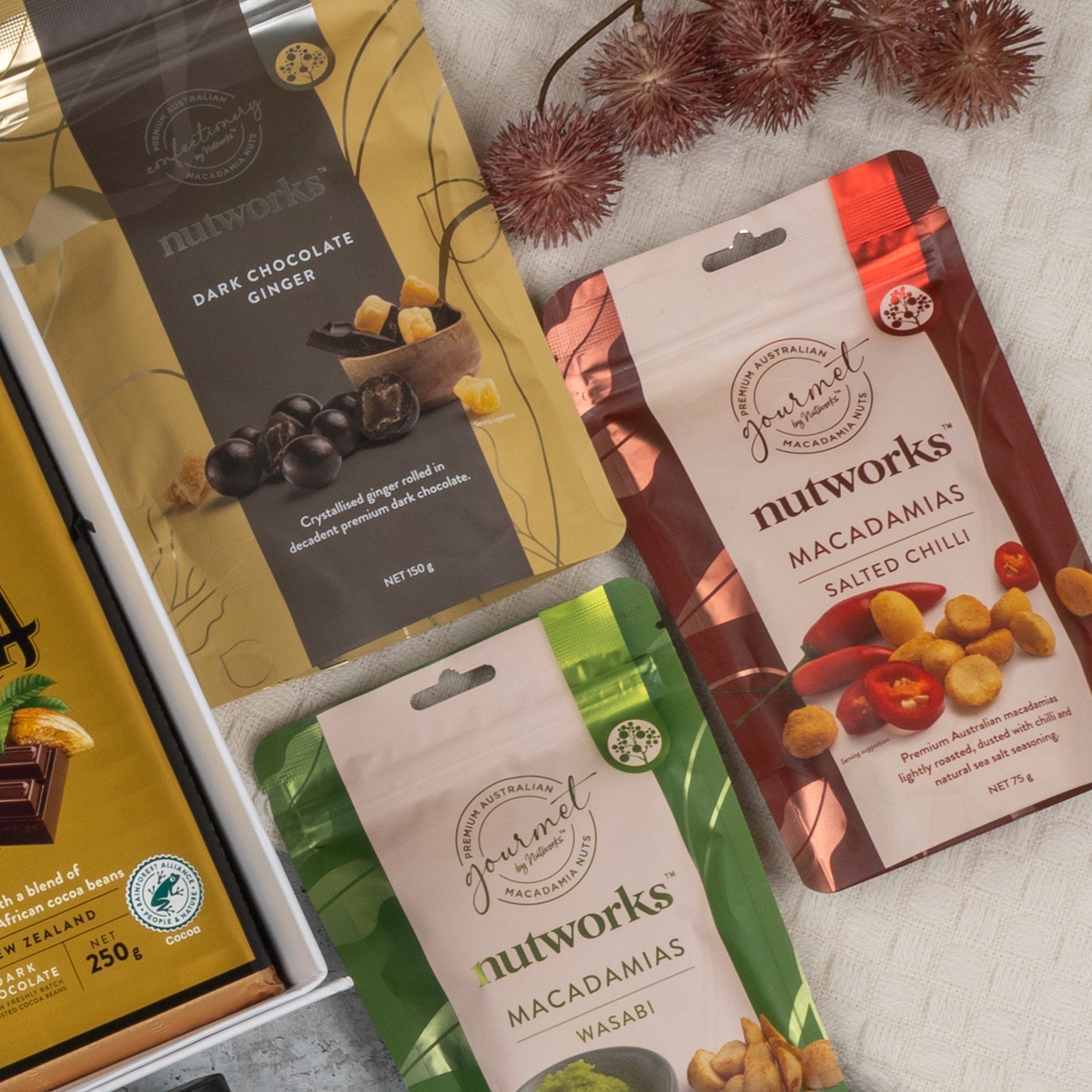 The Hamper Boutique Co’s Ultimate Gourmet Collection Hamper features three Nutworks snack packs—Dark Chocolate Ginger, Macadamias Salted Chilli, and Macadamias Wasabi—arranged on beige cloth with dried flowers and a chocolate box in view.
