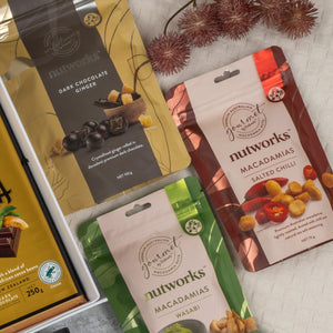 The Hamper Boutique Co’s Ultimate Gourmet Collection Hamper features three Nutworks snack packs—Dark Chocolate Ginger, Macadamias Salted Chilli, and Macadamias Wasabi—arranged on beige cloth with dried flowers and a chocolate box in view.