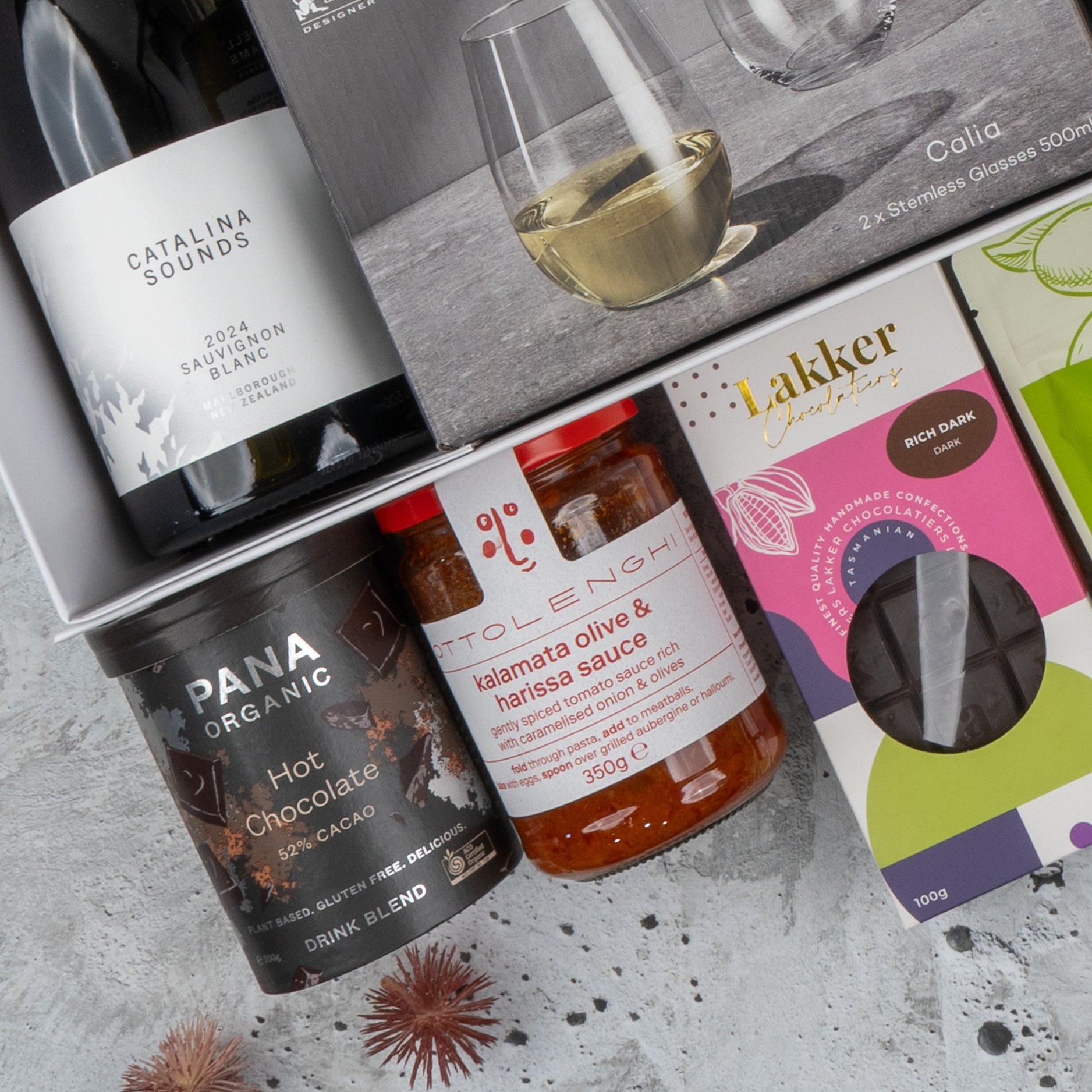 The Hamper Boutique Co’s Ultimate Gourmet Collection Hamper features Catalina Sounds wine, Callia stemless glasses, Pana Organic hot chocolate, Ottolenghi kalamata olive & harissa sauce, and Lakker Chocolates.