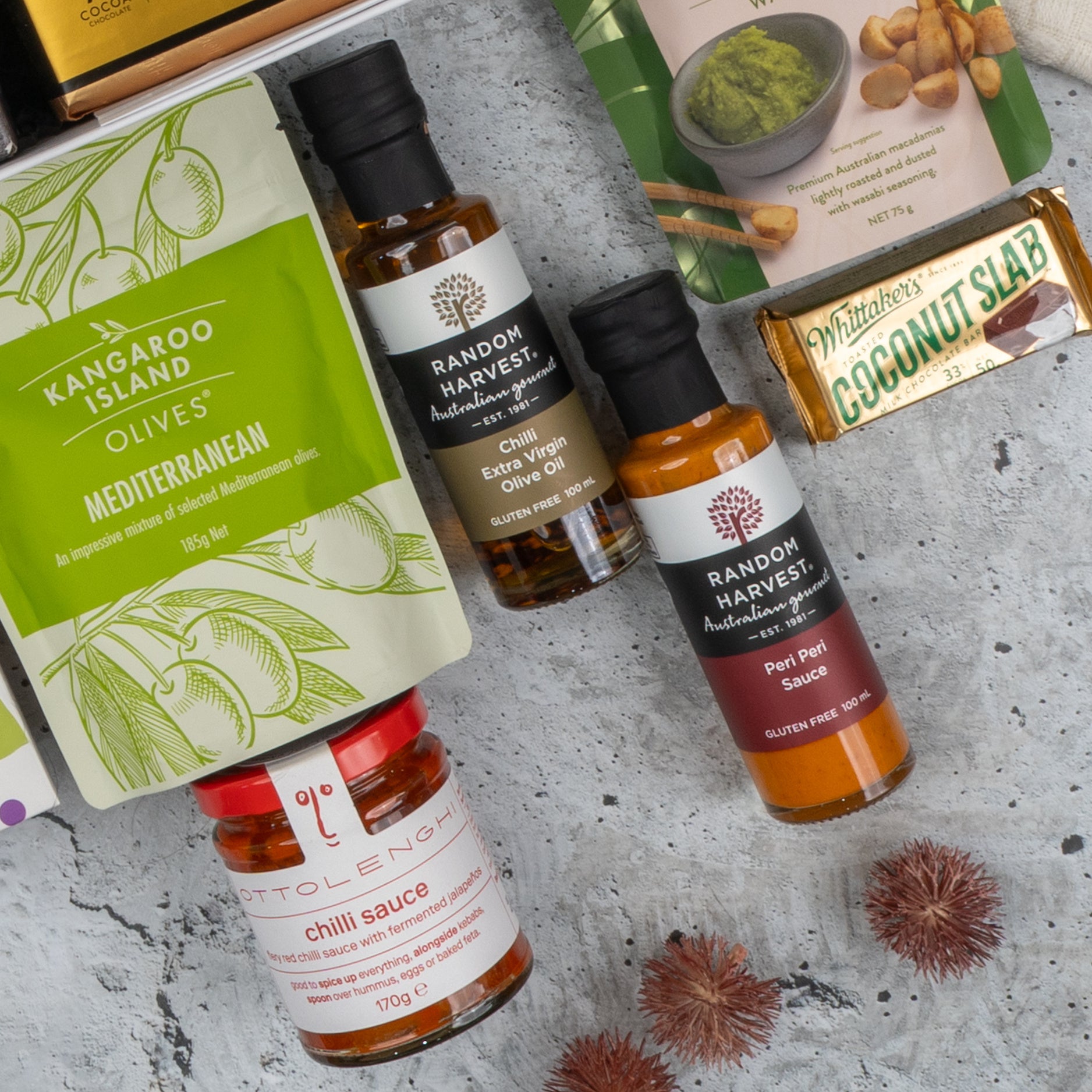 A flat lay of the Ultimate Gourmet Collection Hamper by The Hamper Boutique Co featuring premium olives, gourmet sauces, Whittaker’s Coconut Slab, and a green dip packet on a gray textured surface.