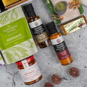 A flat lay of the Ultimate Gourmet Collection Hamper by The Hamper Boutique Co featuring premium olives, gourmet sauces, Whittaker’s Coconut Slab, and a green dip packet on a gray textured surface.